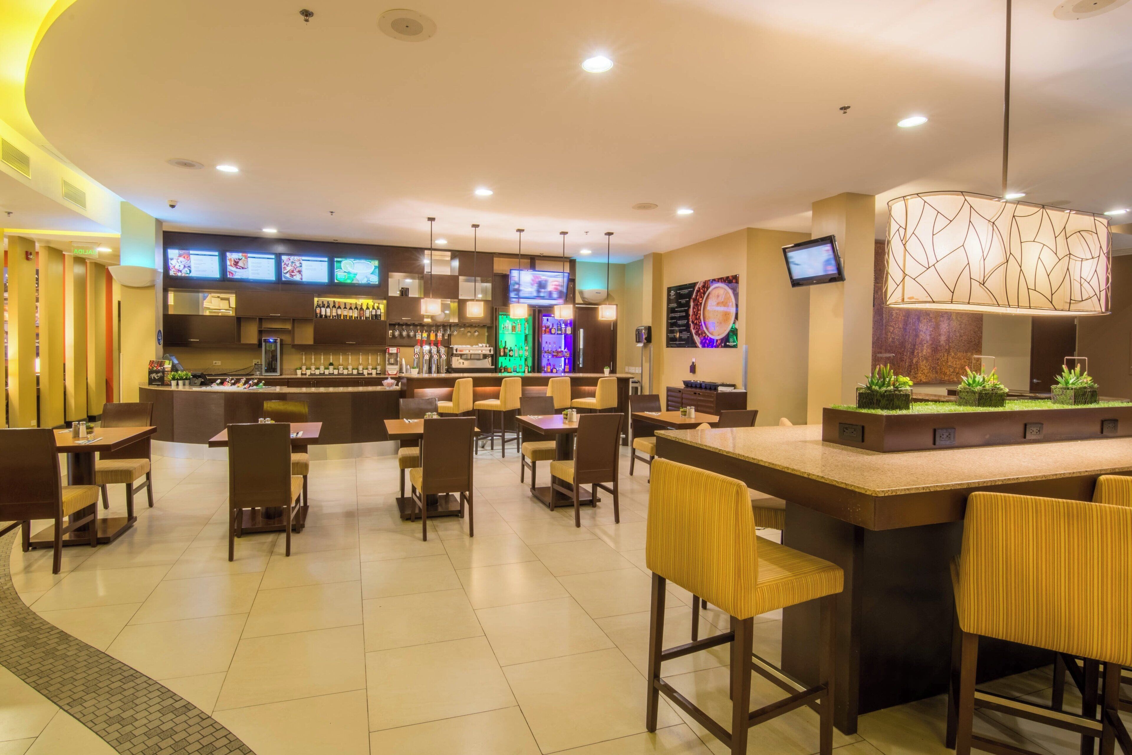 Um lobby moderno de hotel com área de bar, diversos arranjos de assento e um painel digital de cardápio.