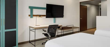 A hotel room with a bed, a desk with a chair, a television mounted on the wall, and a wardrobe.