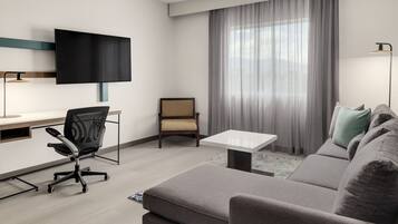 A modern hotel room with a flat-screen TV mounted on the wall, a grey sofa, a small table, a desk with a chair, and a large window with curtains.