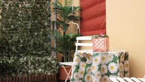 Un pequeño espacio al aire libre con una mesa y sillas cubiertas con un mantel estampado de margaritas, una planta en maceta y un persiano rojo en la pared.