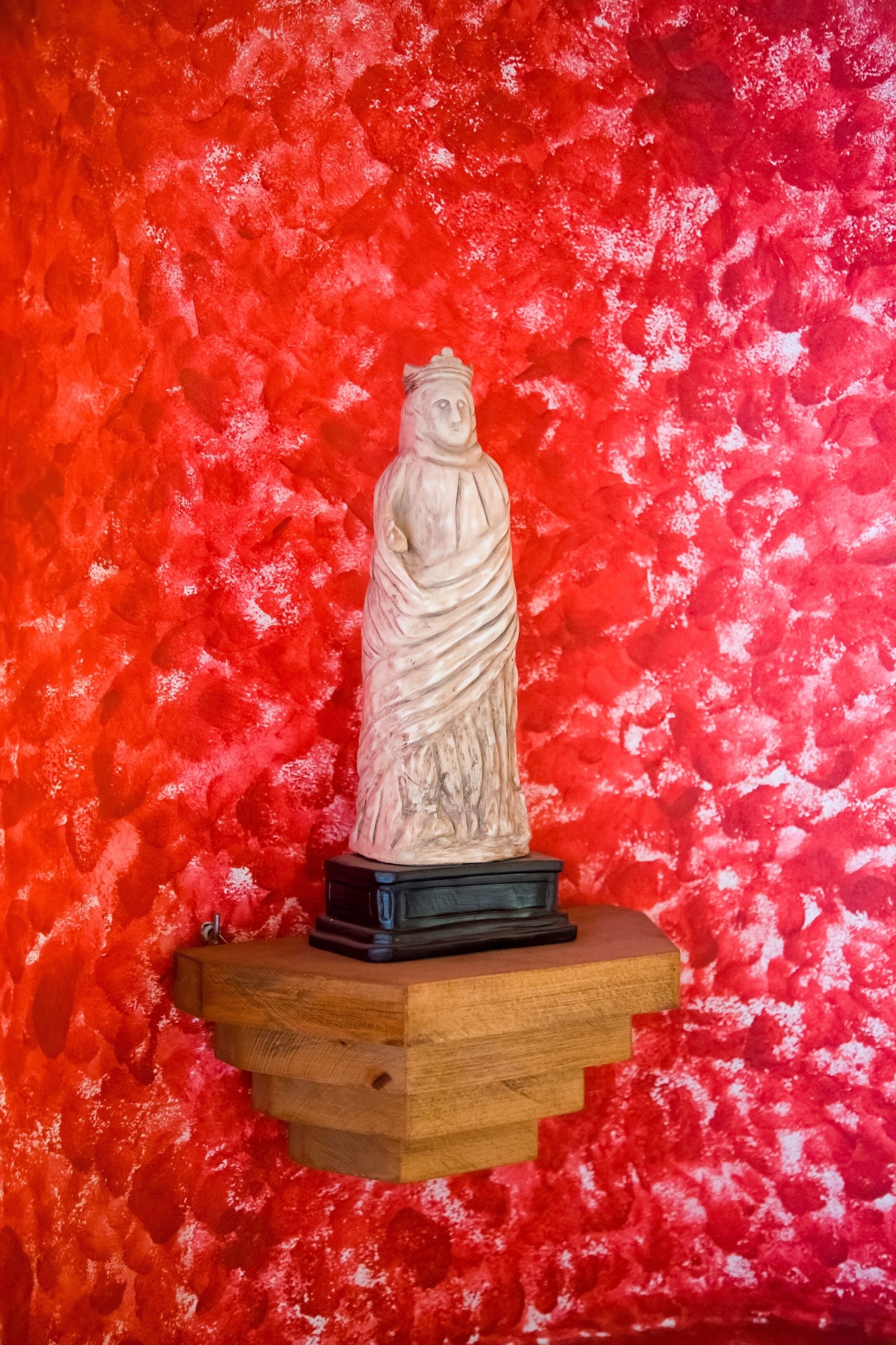 Une statue posée sur une étagère en bois, devant un mur rouge à texture marquée.