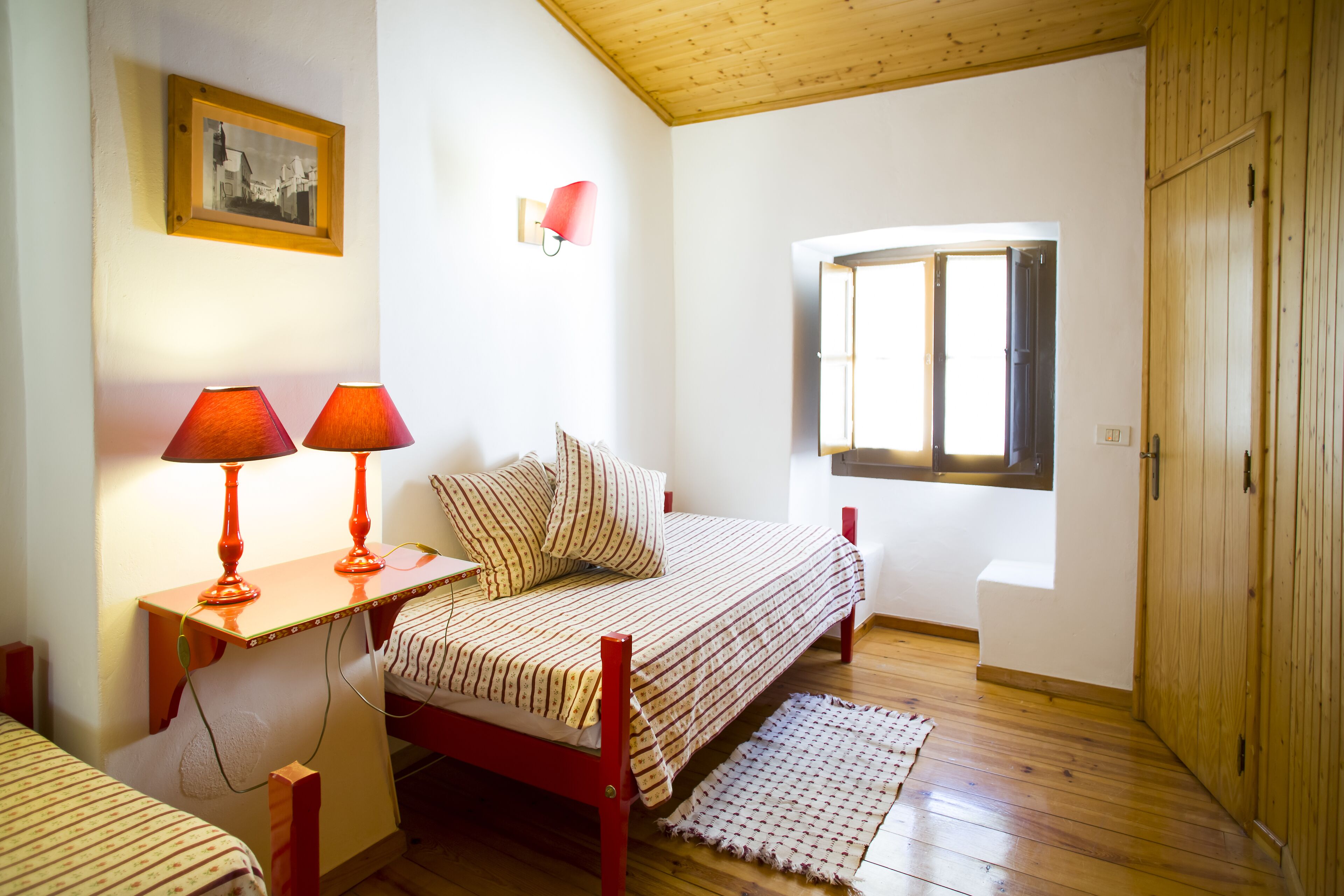 A small, cozy bedroom with a single bed, a nightstand with two lamps, a window with a view, and a wooden wardrobe.