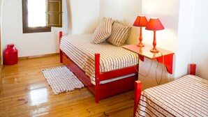 Un dormitorio con techo de madera inclinado, una cama con ropa de cama a rayas, una mesita de noche roja con lámparas, un ventanal pequeño y un cilindro de gas rojo.