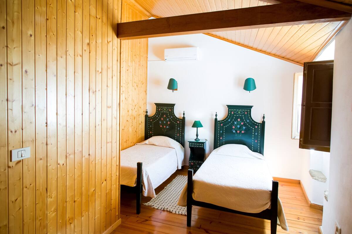 Quarto com duas camas, paredes de madeira, um ventilador de teto e uma pequena mesa com abajur.