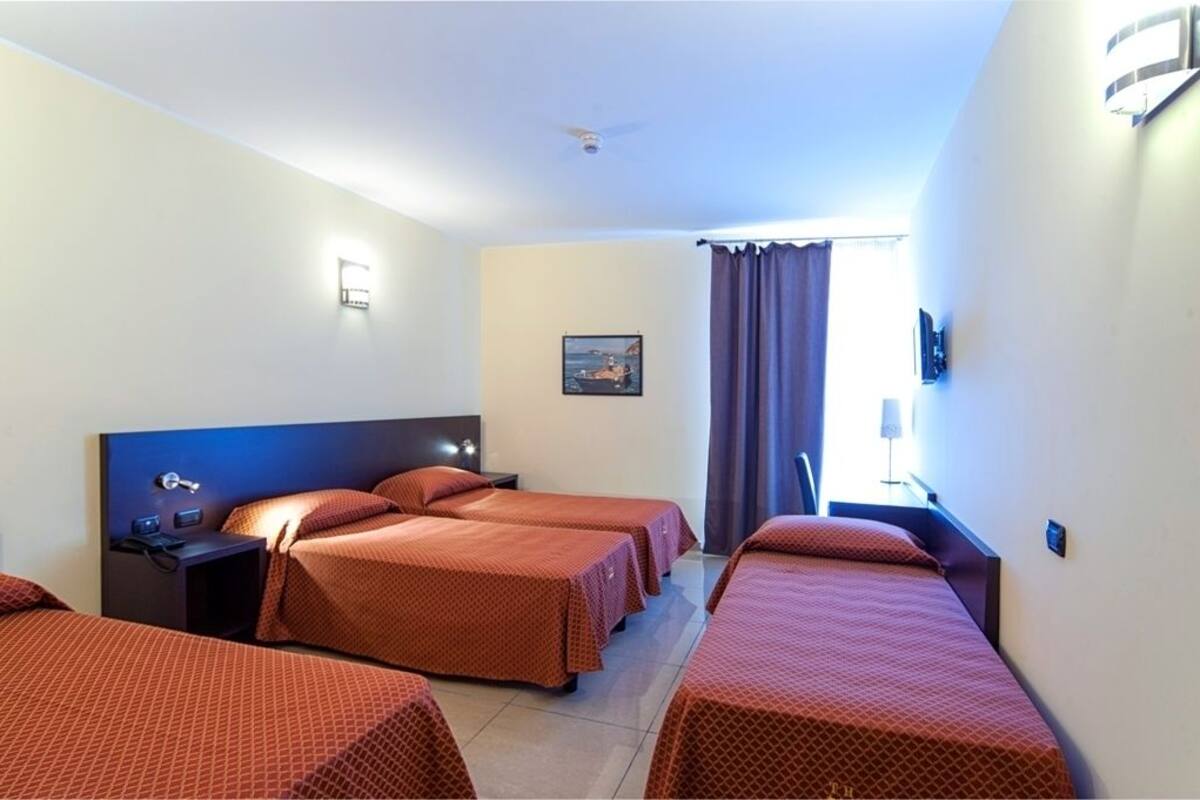A hotel room with four beds arranged in a row, a ceiling fan, a nightstand with a lamp, and a painting on the wall.