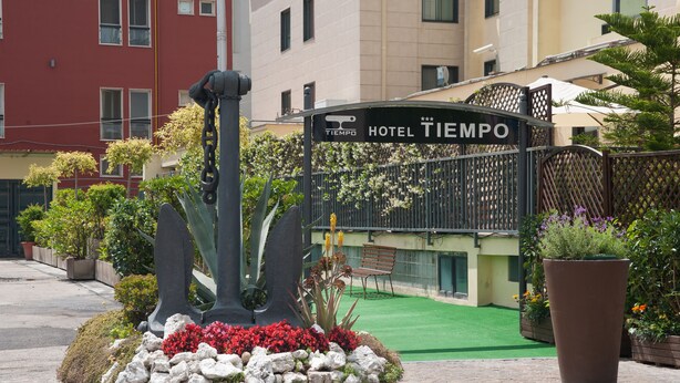 Entrada de un hotel con un cartel que dice "HOTEL TIEMPO", rodeada por un jardín ajardinado con rocas, flores y una jardinera grande.
