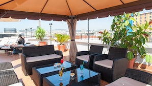 Una terraza en la azotea con mobiliario de mimbre, plantas en macetas y una vista despejada del paisaje urbano.