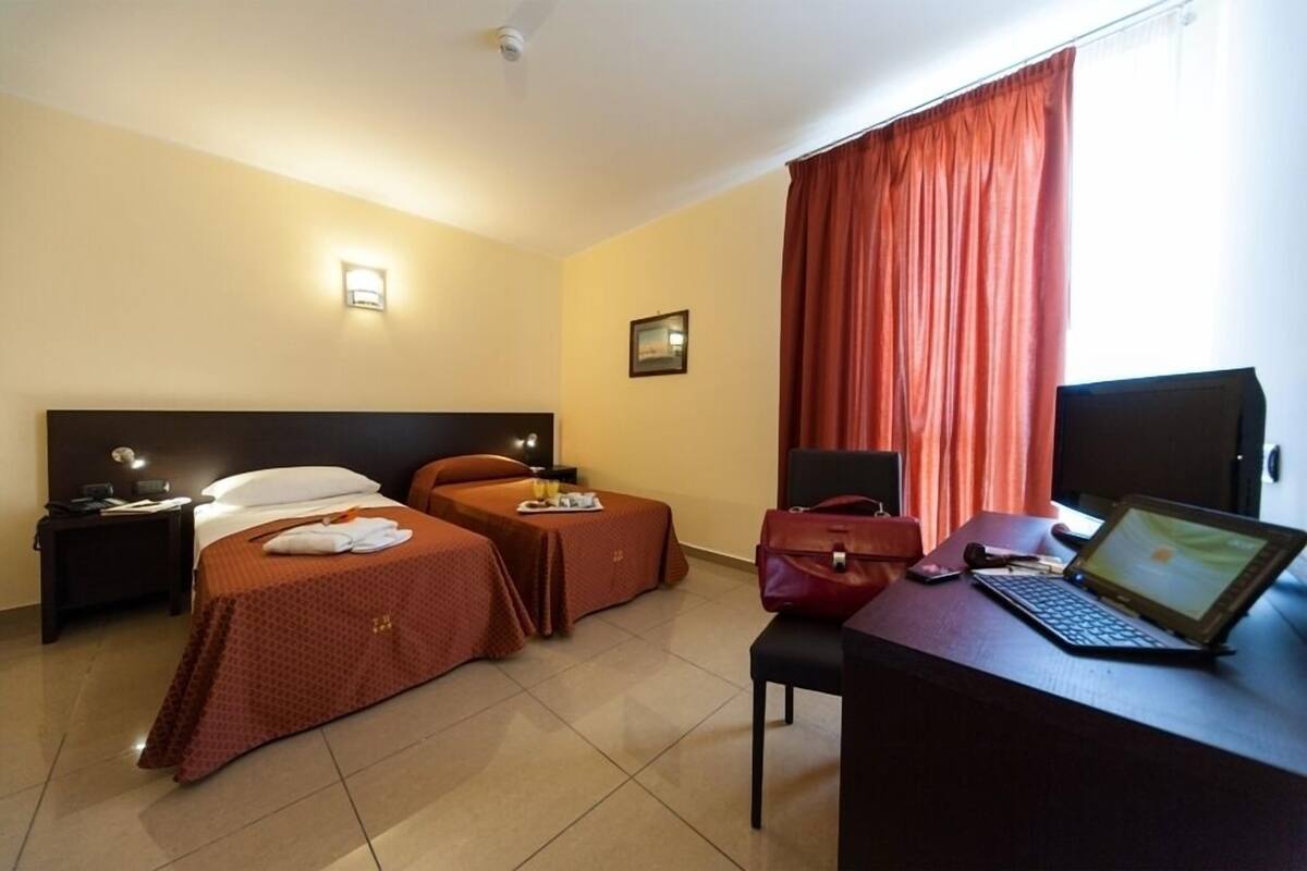 A hotel room with two beds, a desk with a laptop, a chair, a desk lamp, a nightstand, and a window with curtains.