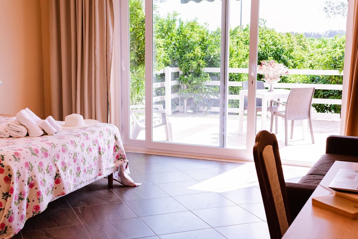 Habitación de hotel con cama, una silla, una mesa y vistas a un patio con mobiliario exterior y vegetación.