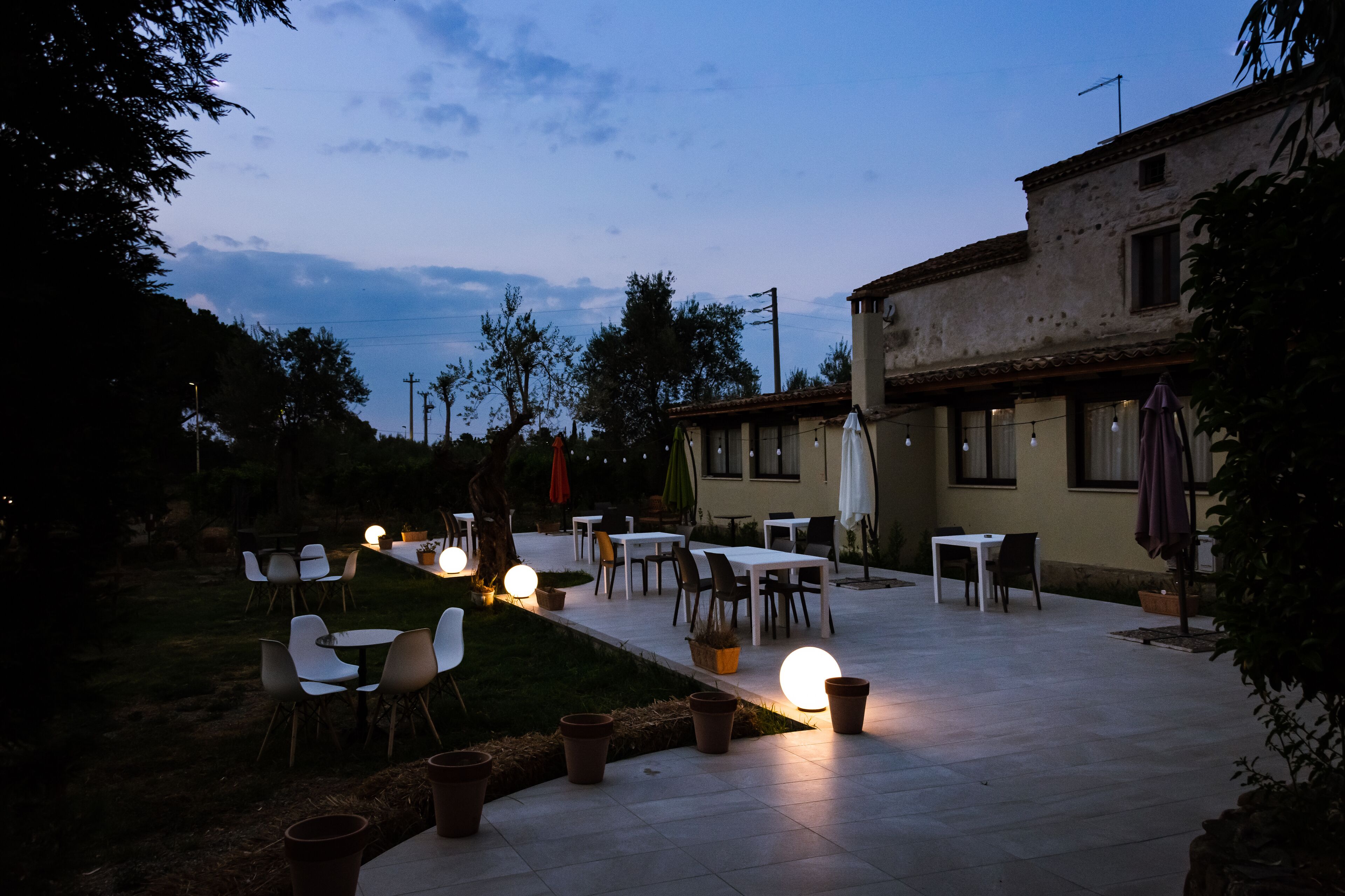 Un patio al aire libre con mesas y sillas, rodeado de macetas con plantas y un edificio.