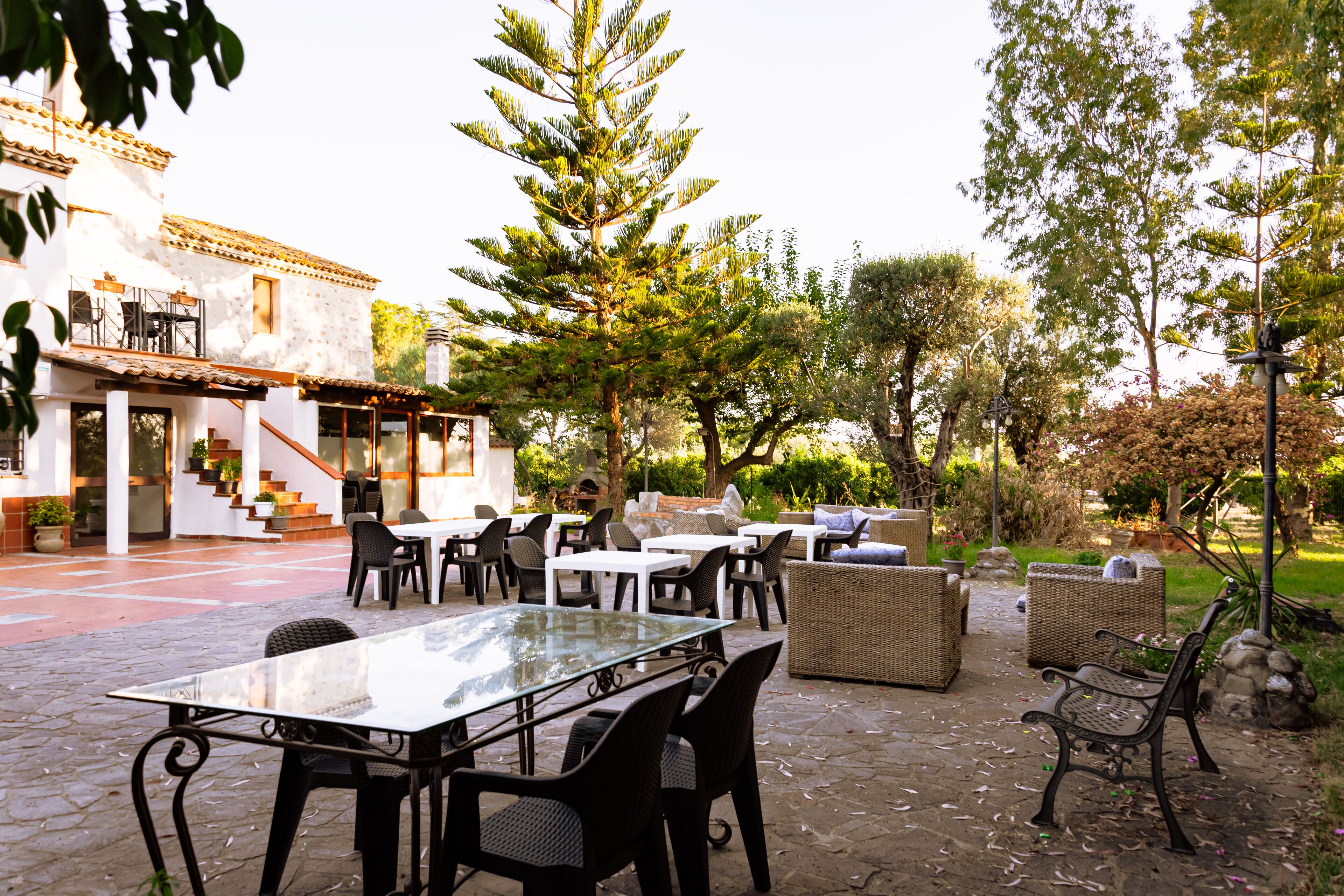Un patio al aire libre con mesas y sillas, una mesa con cubierta de vidrio, mobiliario de mimbre y una escalera que lleva a un nivel superior.