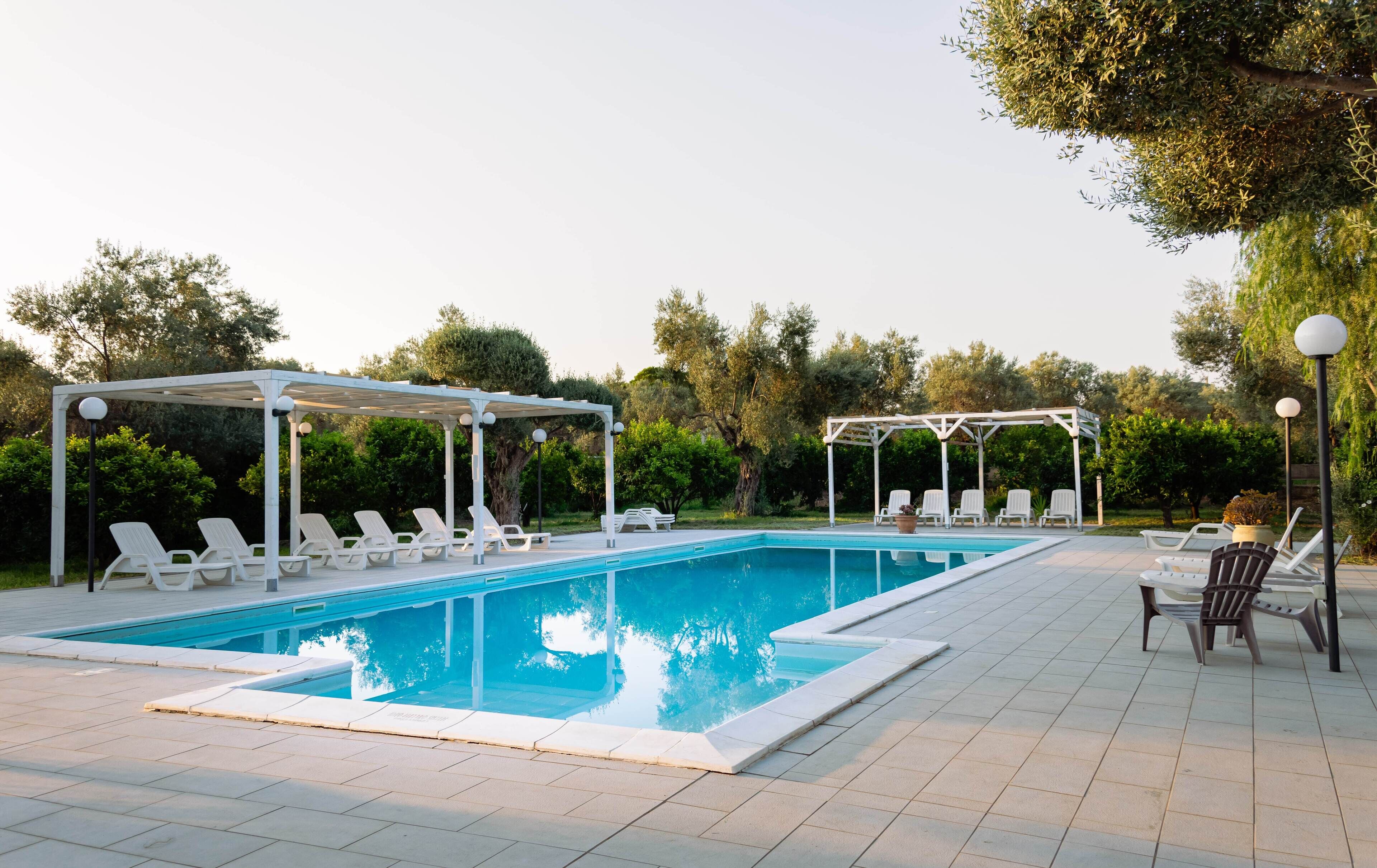 Piscina con sillas reclinables blancas y pérgolas, rodeada de vegetación.