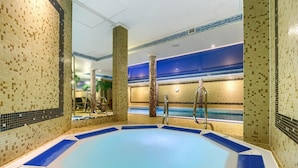 Een groot binnenzwembad met betegelde vloer en wanden, omgeven door kolommen en een blauw verlichte muur.