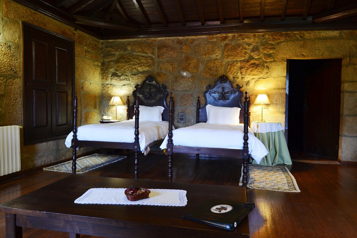 Una habitación con dos camas de madera, paredes de piedra y una mesa de madera con una cesta y un libro.
