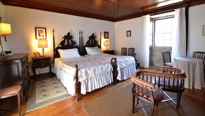 A traditional room with a wooden ceiling, a large bed with floral bedding, a wooden chair, a small table, and a desk with a lamp. - Casa dos Assentos de Quintiães (Barcelos)