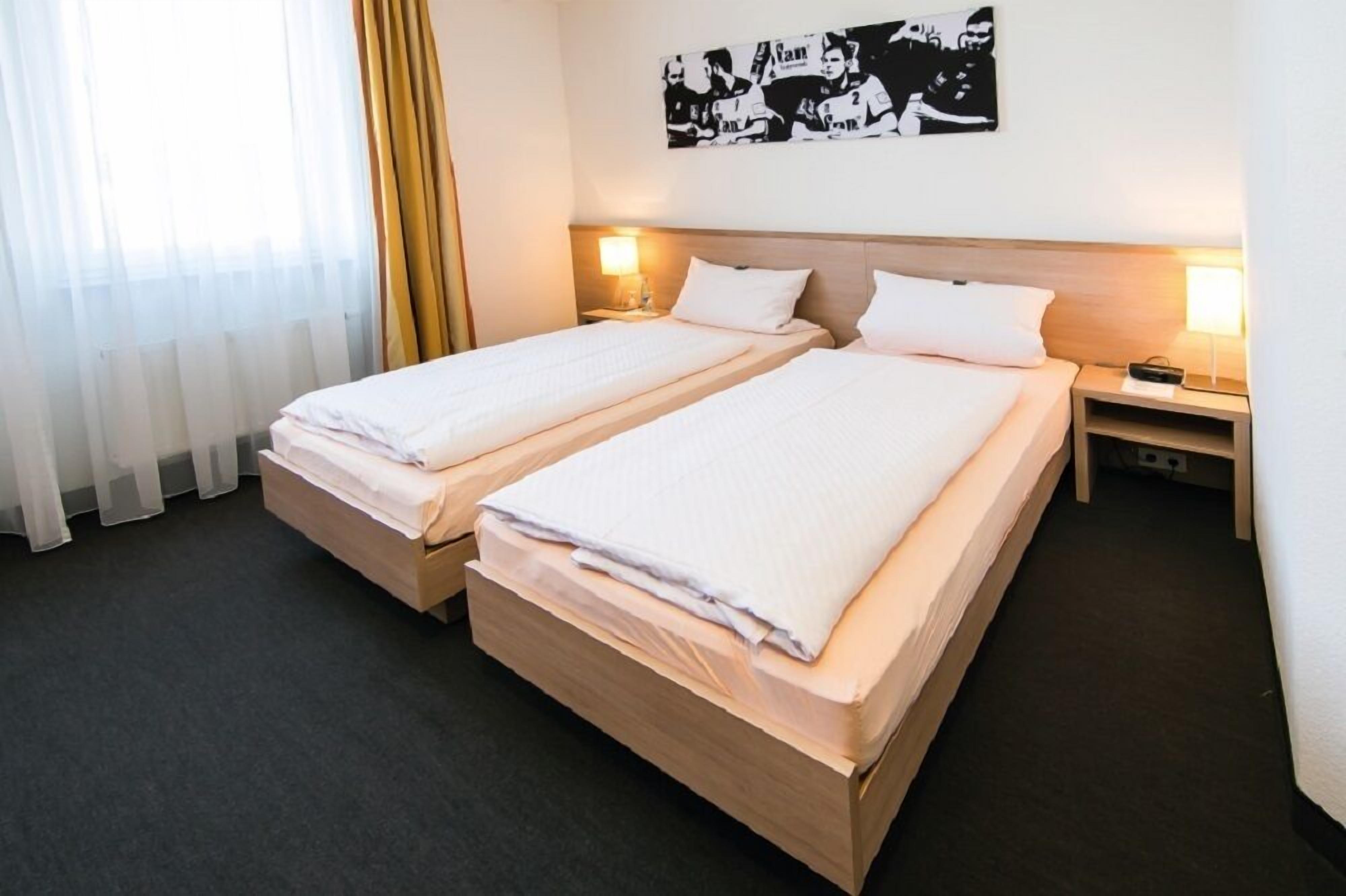 A hotel room with two single beds, a wooden headboard, bedside lamps, and a large window with sheer curtains.