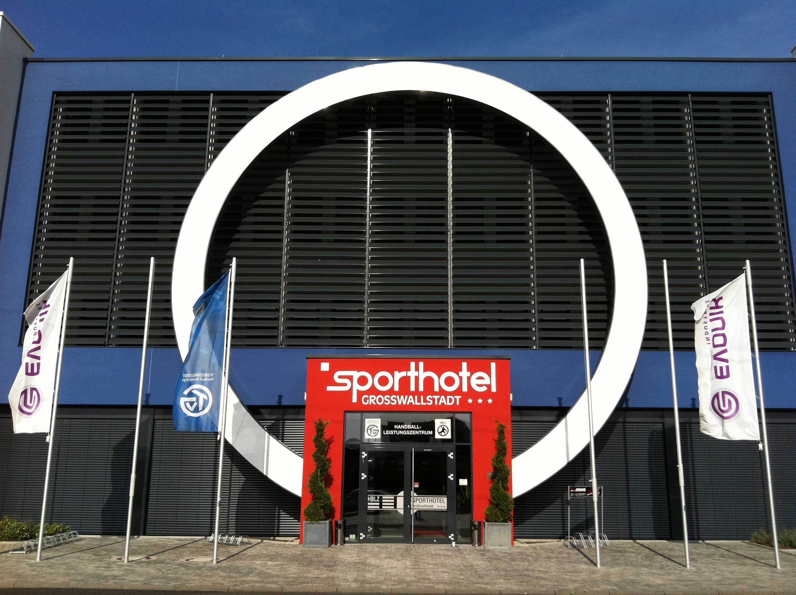 The entrance of Sporthotel Grosswallstadt with a large circular archway, flags, and a sign indicating a handball and sports center.