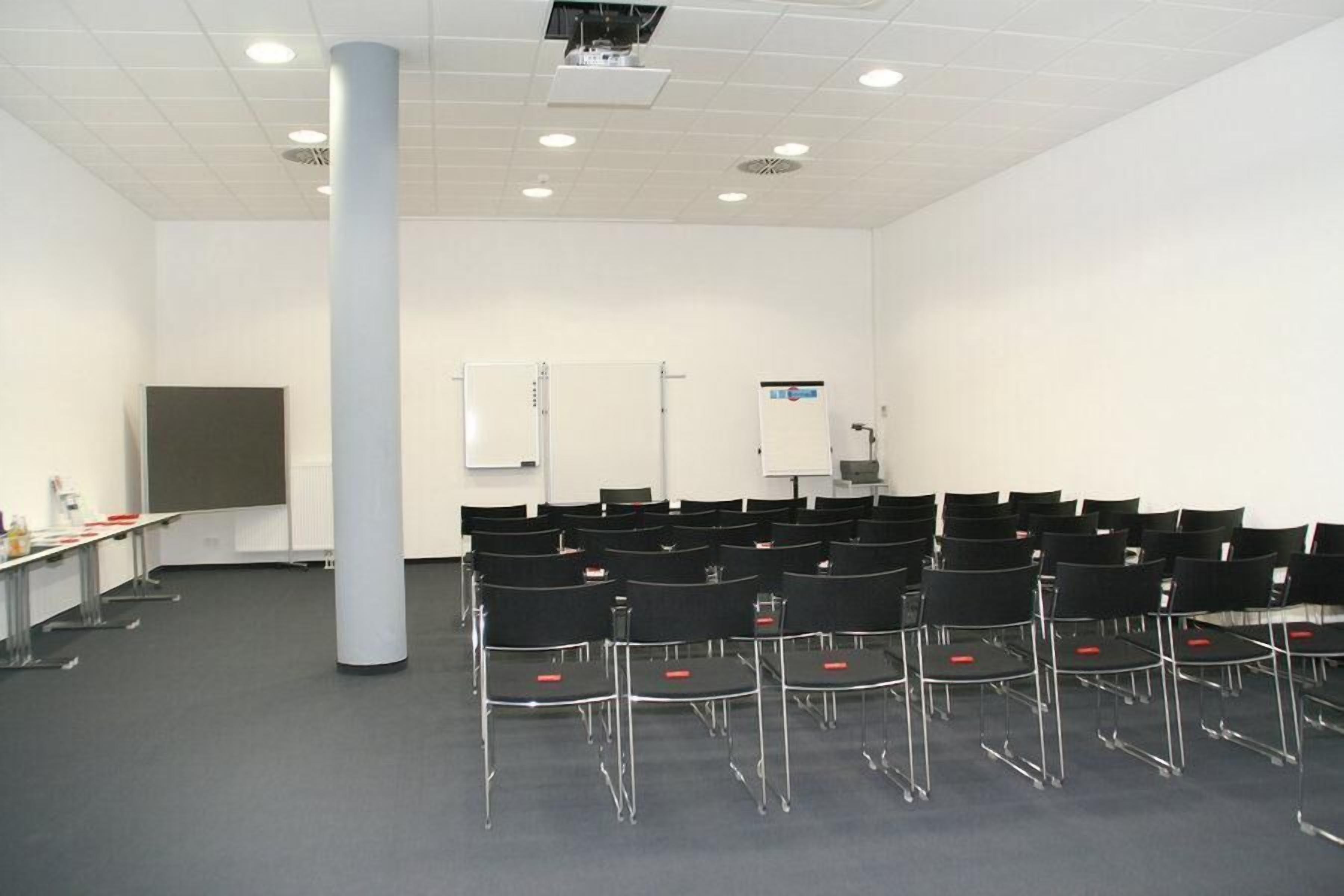Un salón de conferencias con filas de sillas, un proyector, una pizarra y una pantalla para presentaciones.