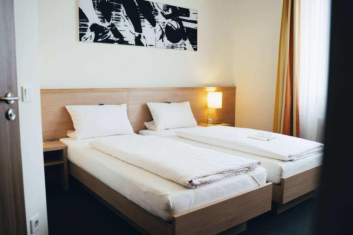 A hotel room with two single beds, a bedside table with a lamp, and a large abstract painting on the wall.