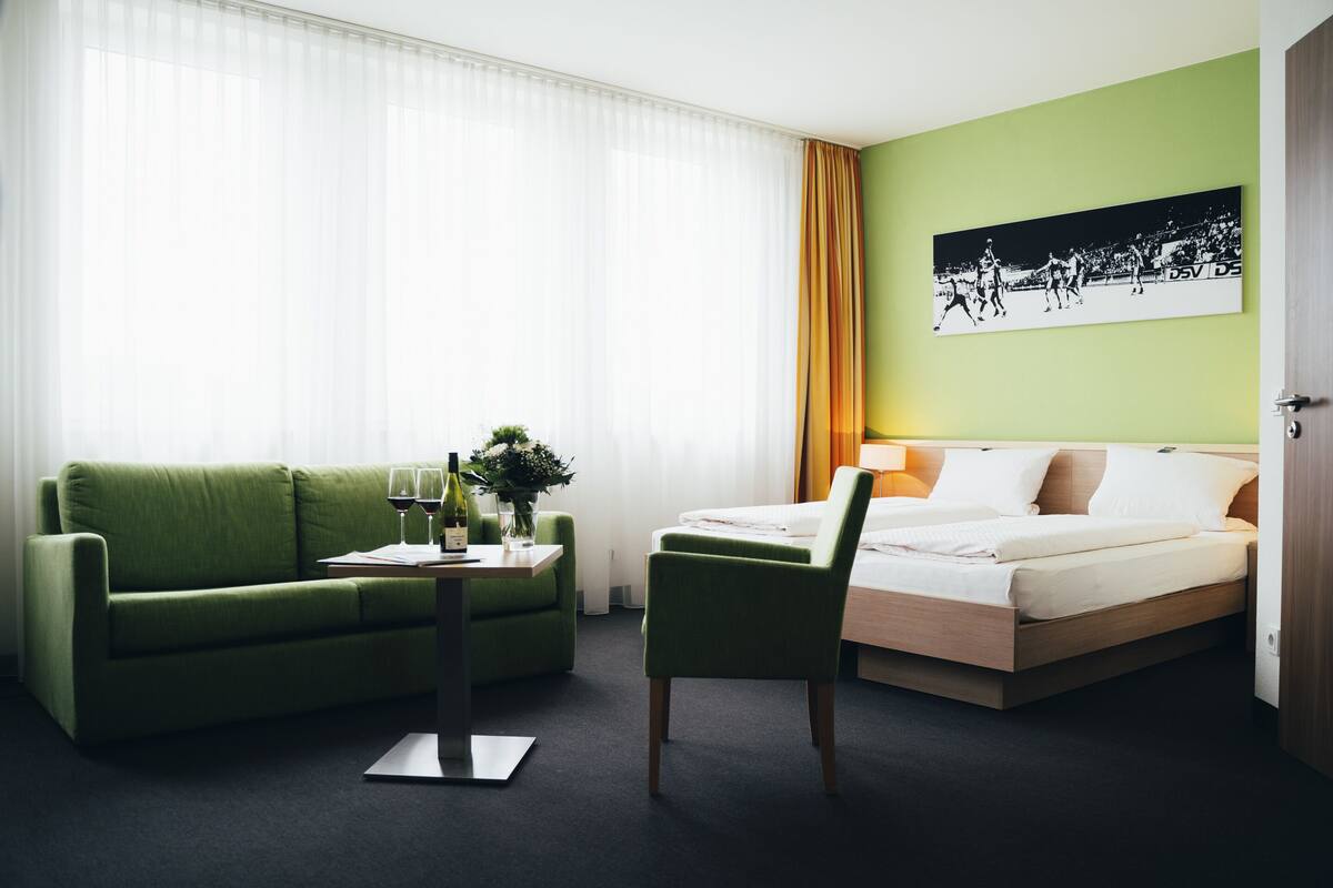 A hotel room with a green sofa, a bed, a desk with a lamp, and a framed picture on the wall.