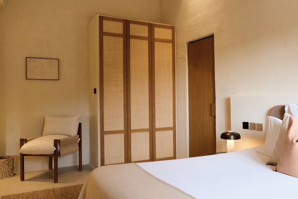 A hotel room with a bed, a chair, a wardrobe, and a bedside table with a lamp.