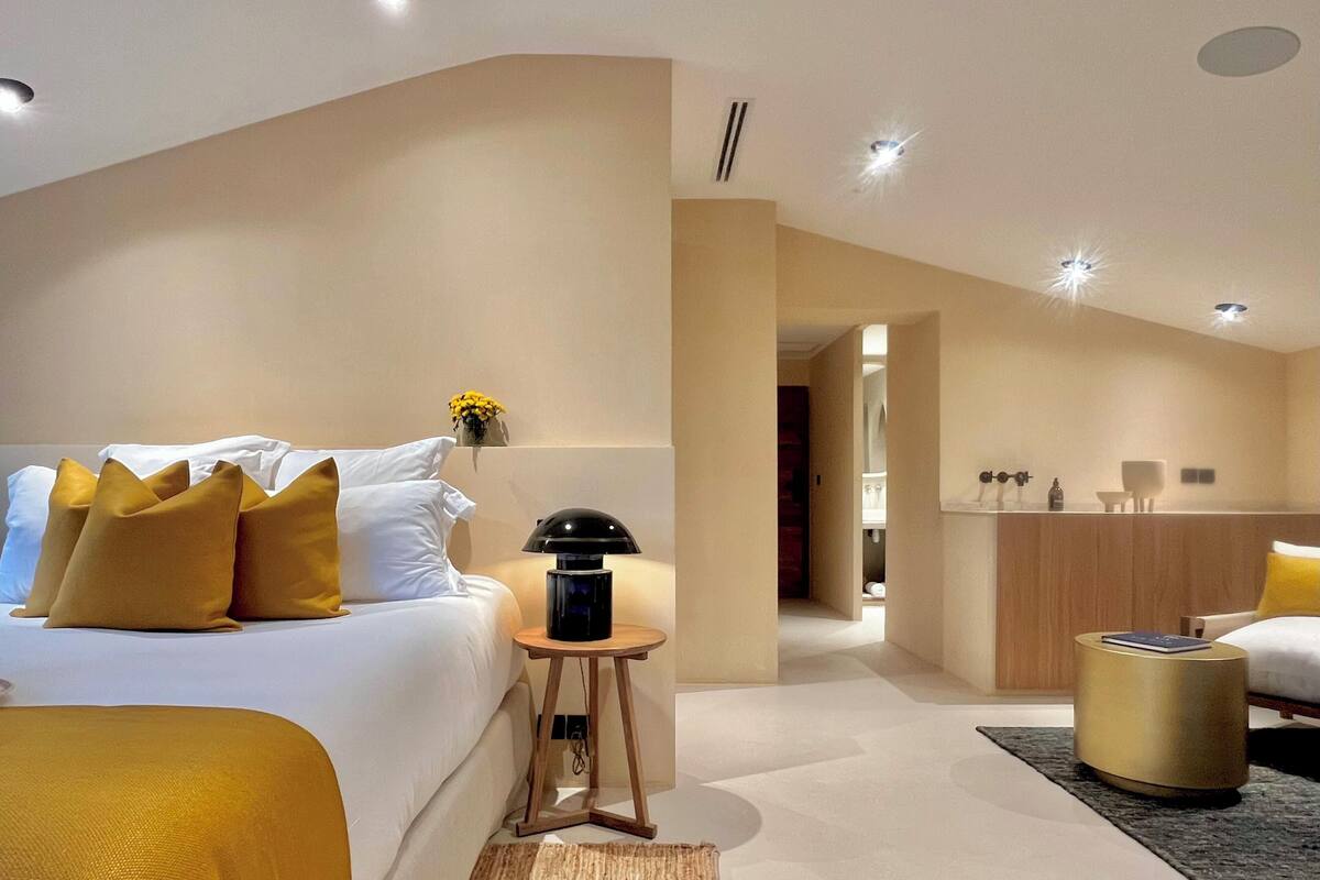 A modern hotel room with a large bed, a bedside table with a lamp, a small round table, and a bathroom visible through an open door.