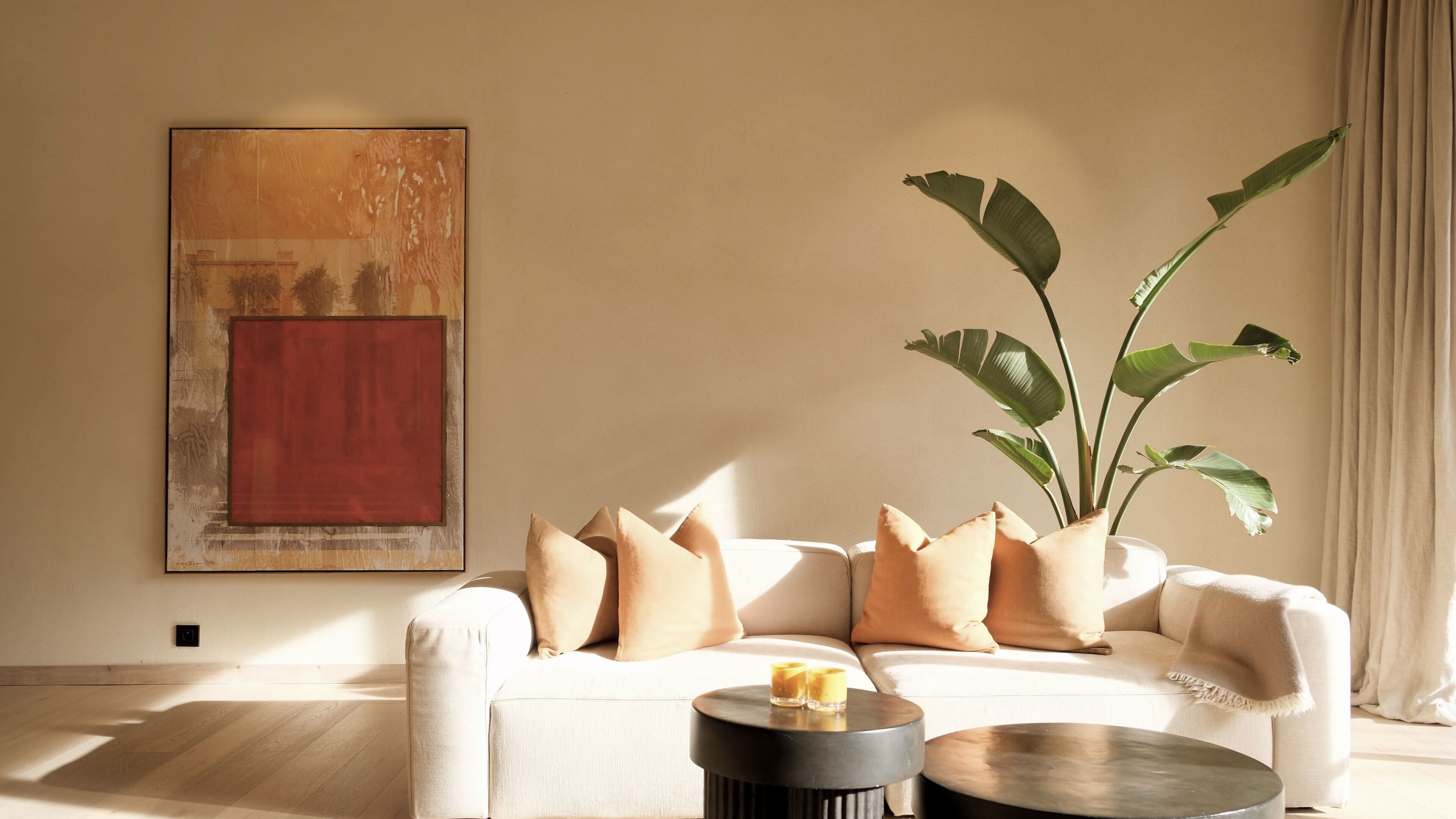 A living room with a beige sofa, a large abstract painting, a potted plant, and a coffee table with two candles.