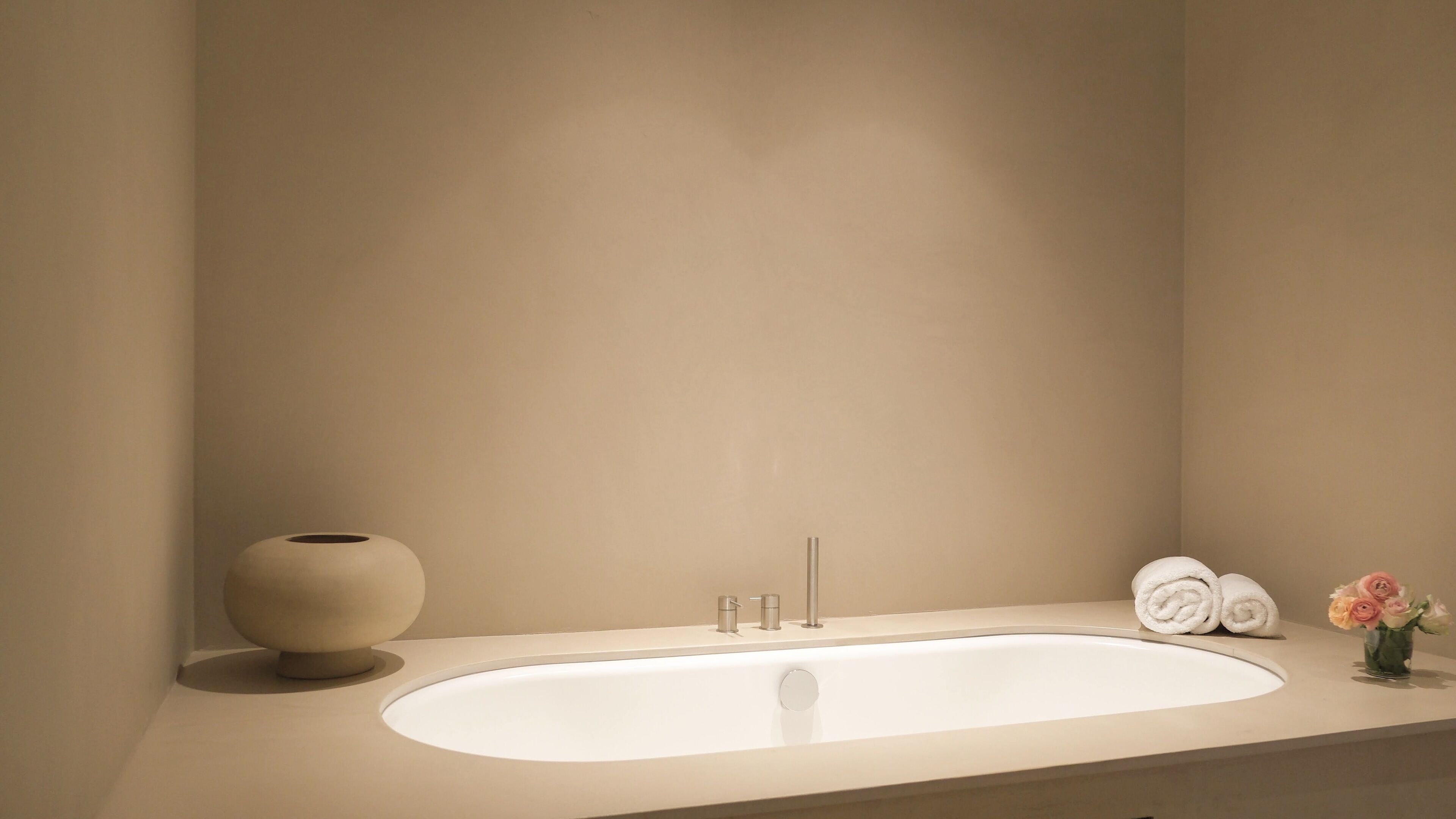 A modern bathroom with a large bathtub, a round vase, and neatly rolled towels.