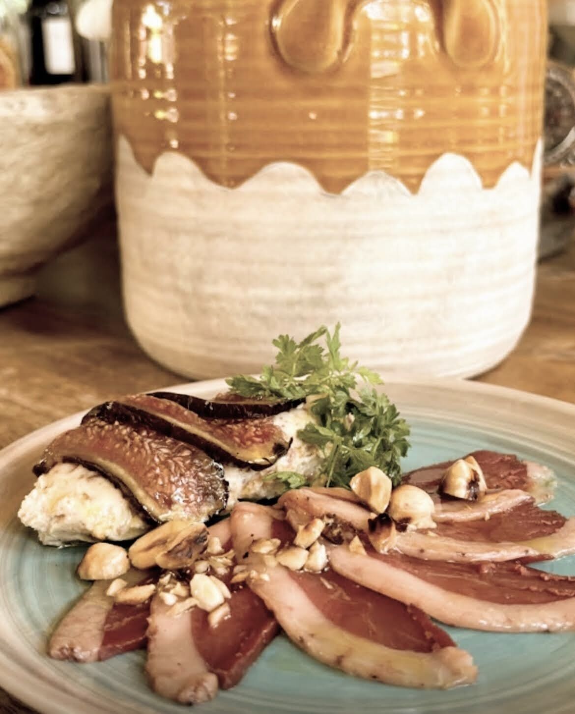 Une assiette de charcuteries et de champignons, garnie de persil, est posée sur une table. On aperçoit en arrière-plan un pot en céramique.