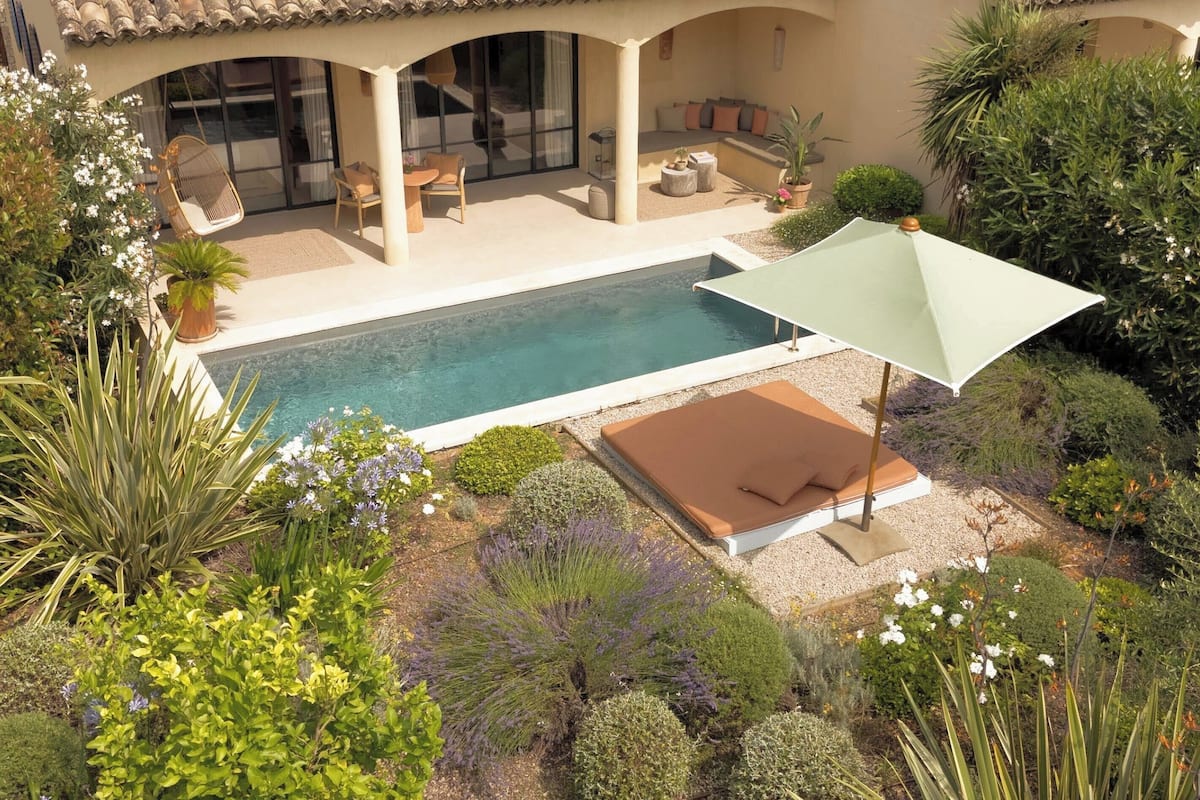A pool area with a lounge chair and umbrella, surrounded by various plants and a tiled roof structure.
