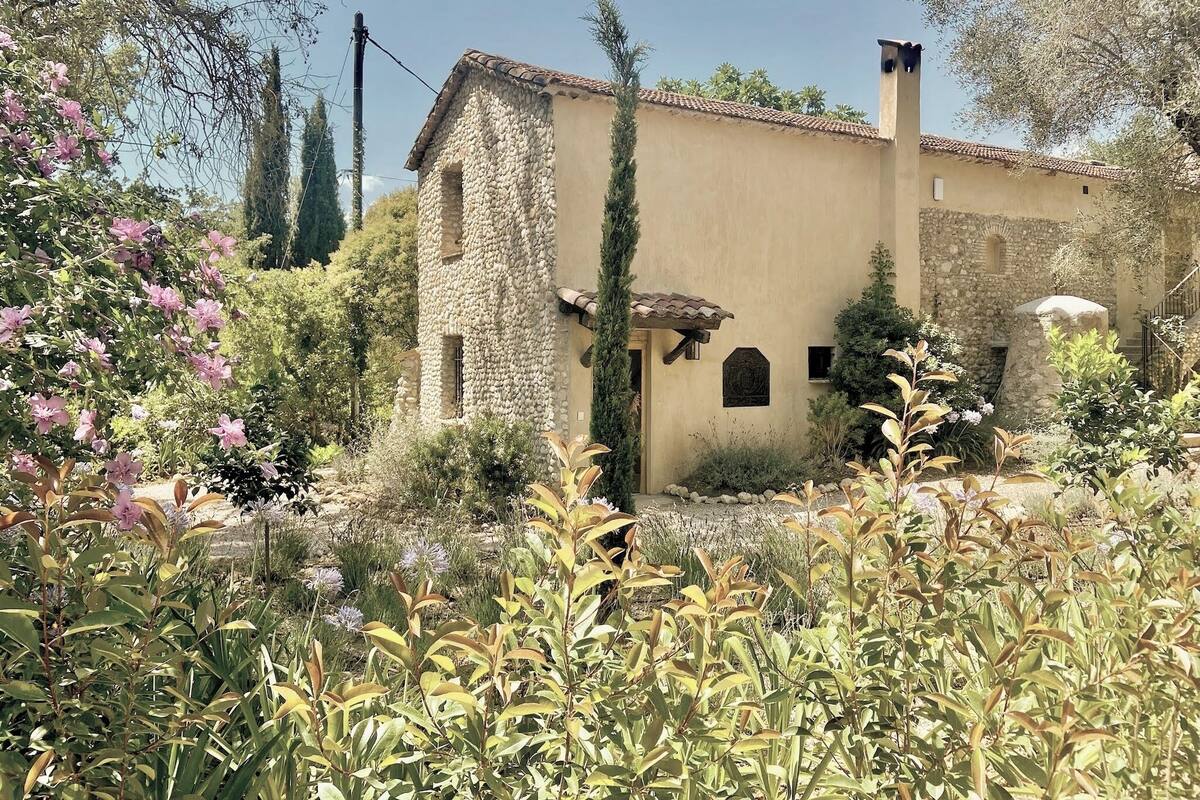 A rustic stone building with a tiled roof, surrounded by lush greenery and flowering plants.
