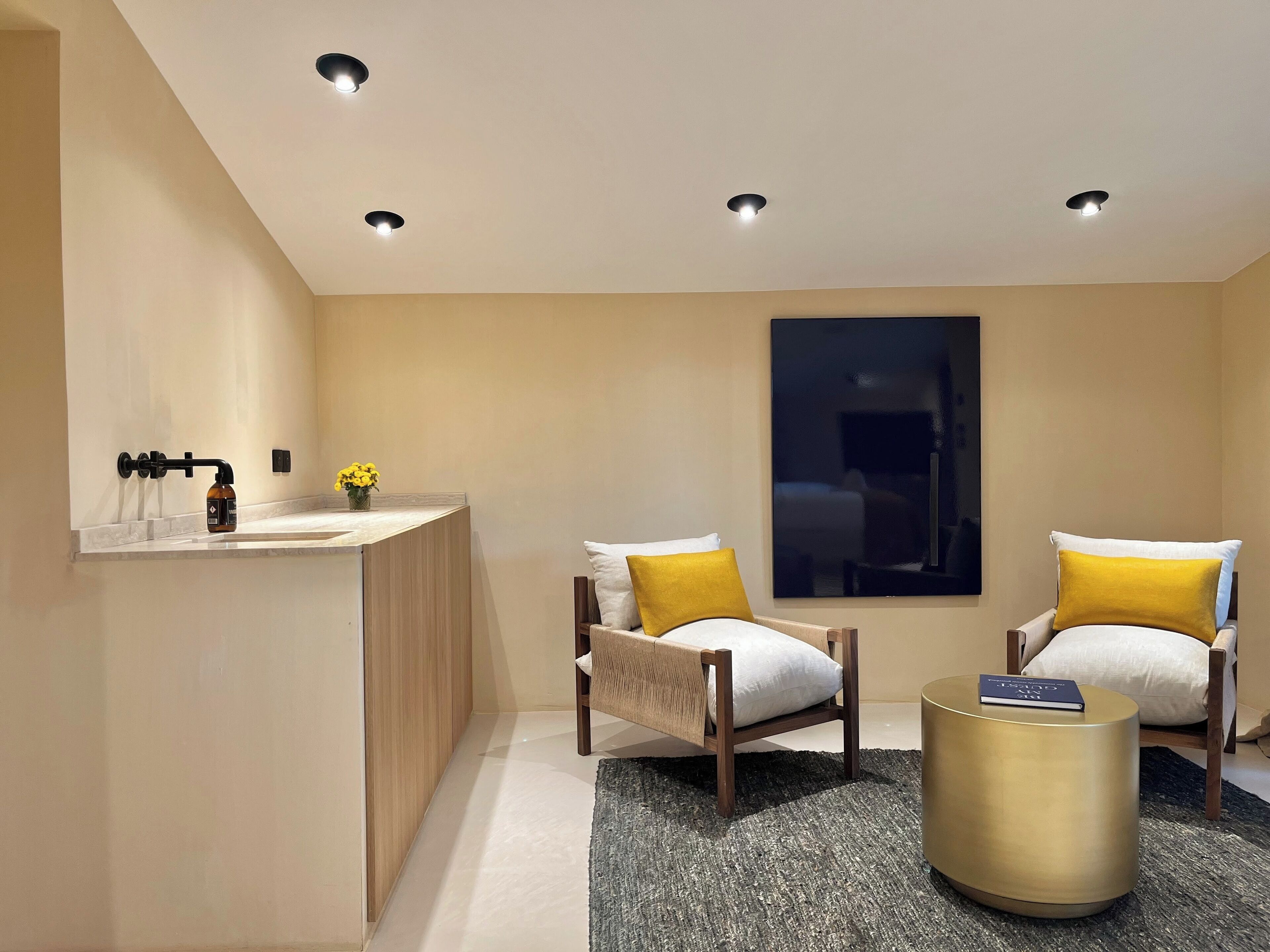 A modern bathroom with a vanity, two chairs, a round gold side table, and recessed ceiling lights.