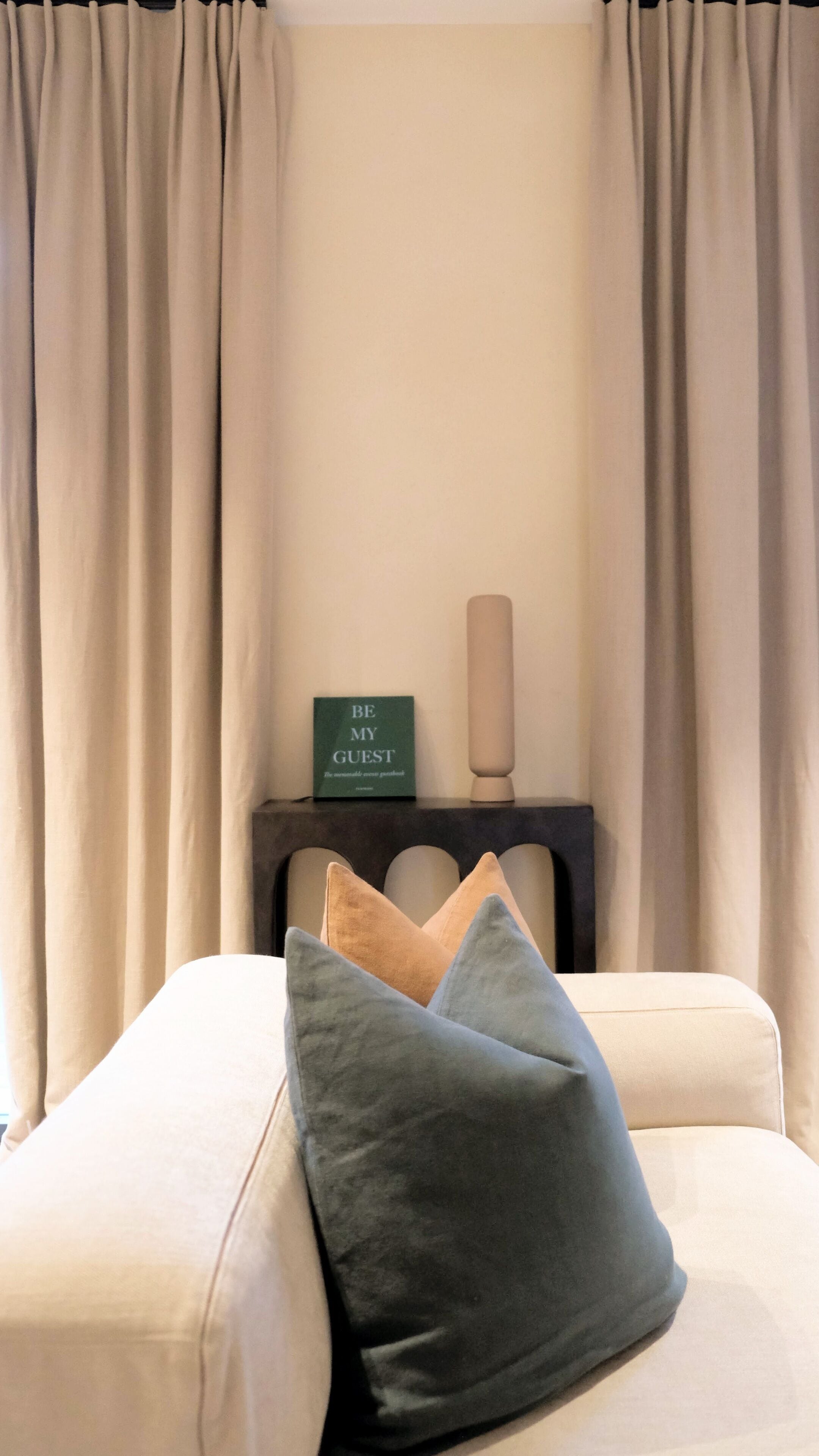 A bedroom with beige curtains, a bedside table with a green sign, and two pillows.