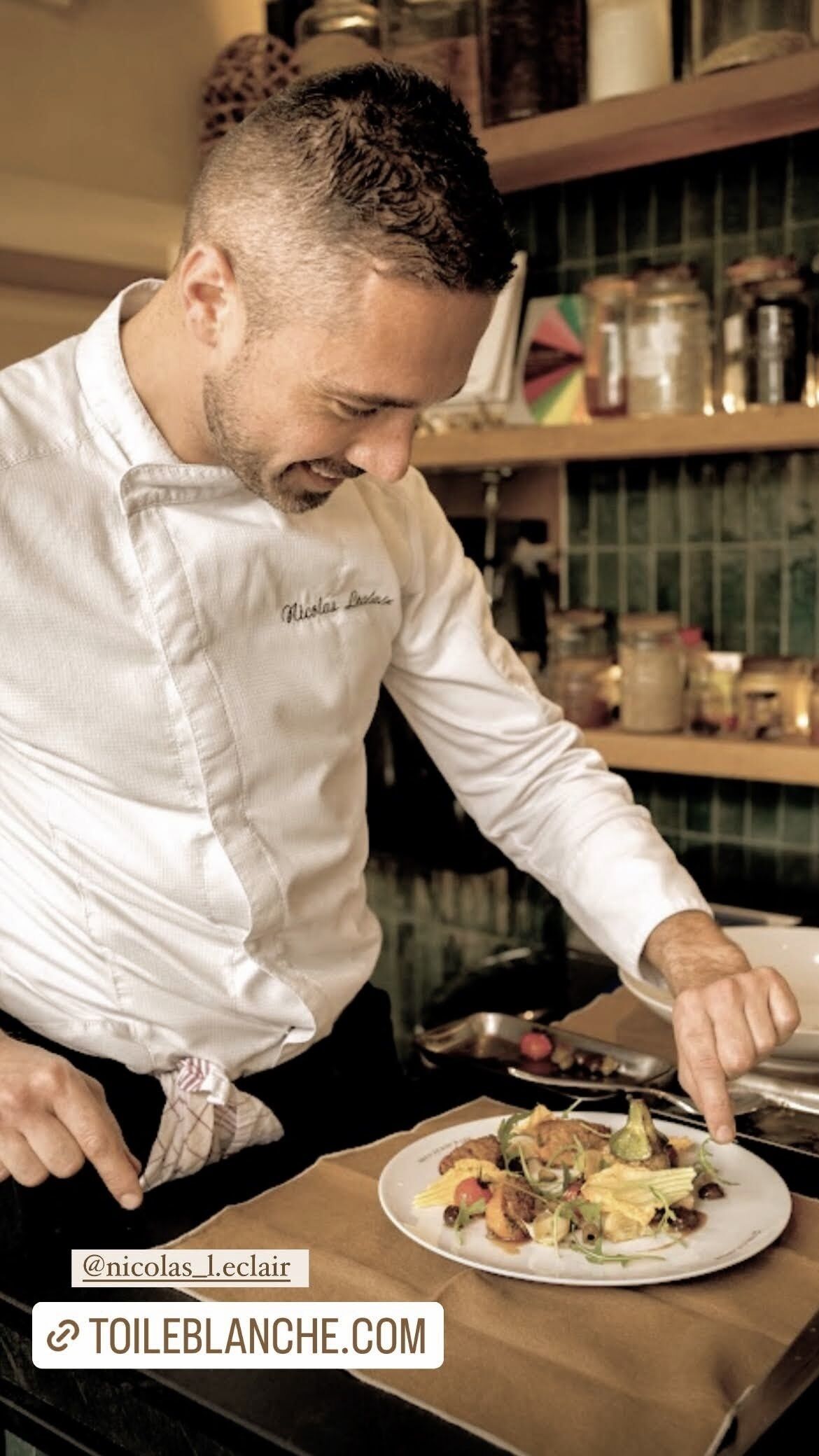 A chef in a white uniform with a name tag, serving a plate of food in a kitchen setting.