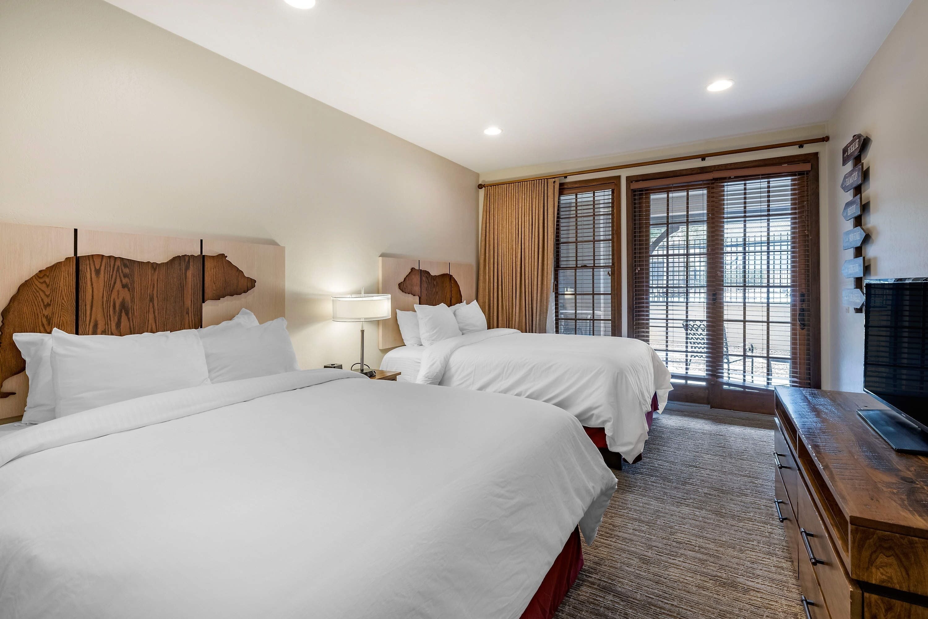 A hotel room with two beds, a wooden headboard, a TV, and a large window with blinds.