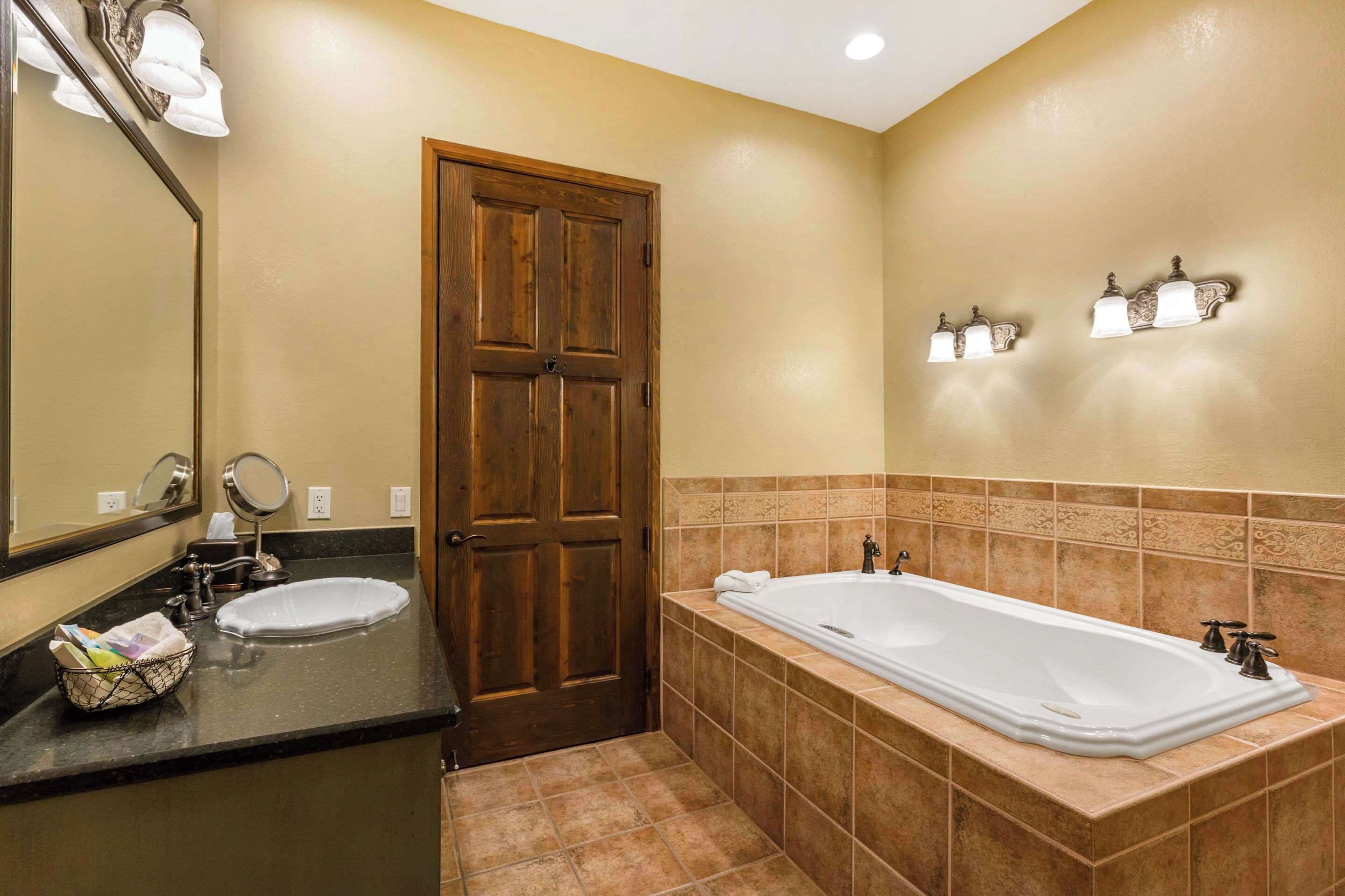 A bathroom with a wooden door, two sinks, a bathtub, and wall-mounted lights.