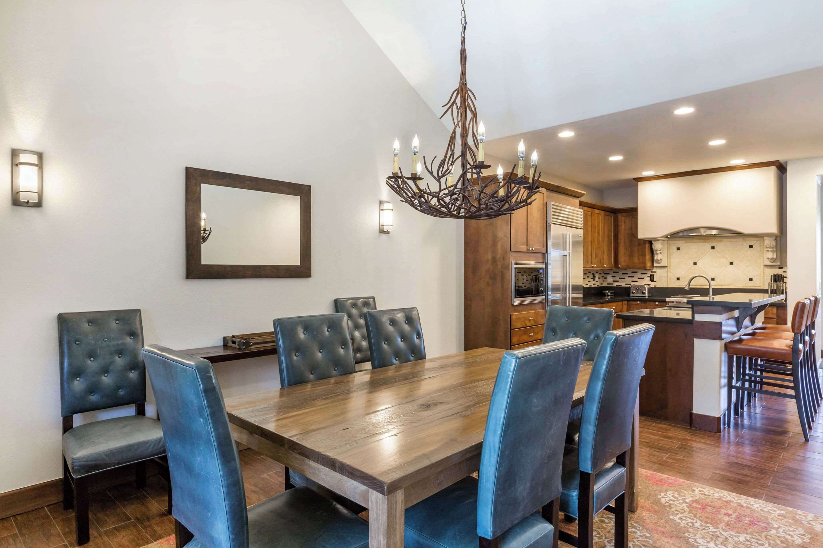Un comedor con mesa de madera y sillas tapizadas en azul, una araña y una cocina al fondo.