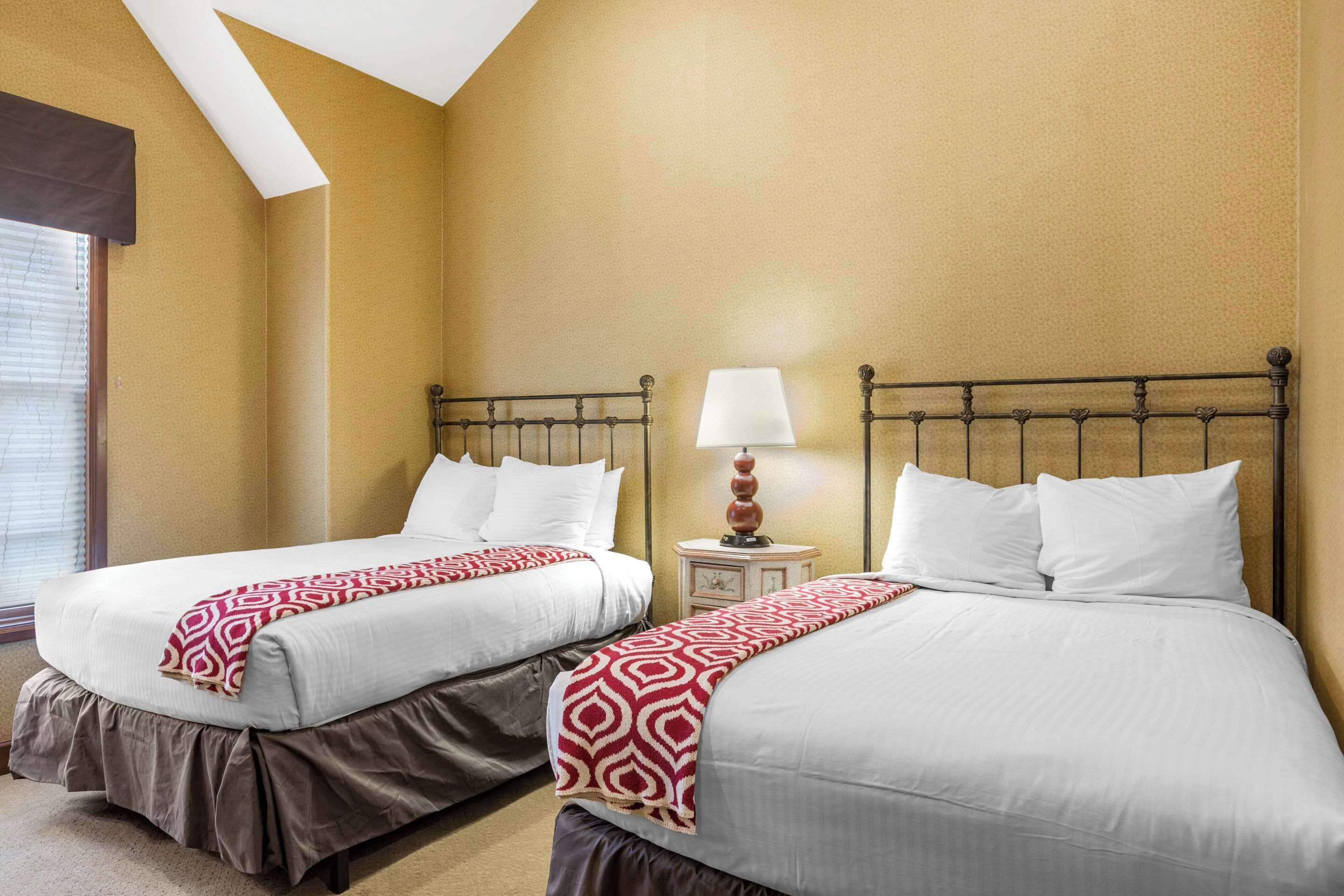 Une chambre d’hôtel avec deux lits, une table de chevet avec une lampe et une fenêtre avec des stores.