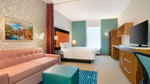 A hotel room with a bed, a sofa, a desk, a TV, and a painting on the wall. - Home2 Suites by Hilton Charlotte I-77 Tyvola Rd (Charlotte)
