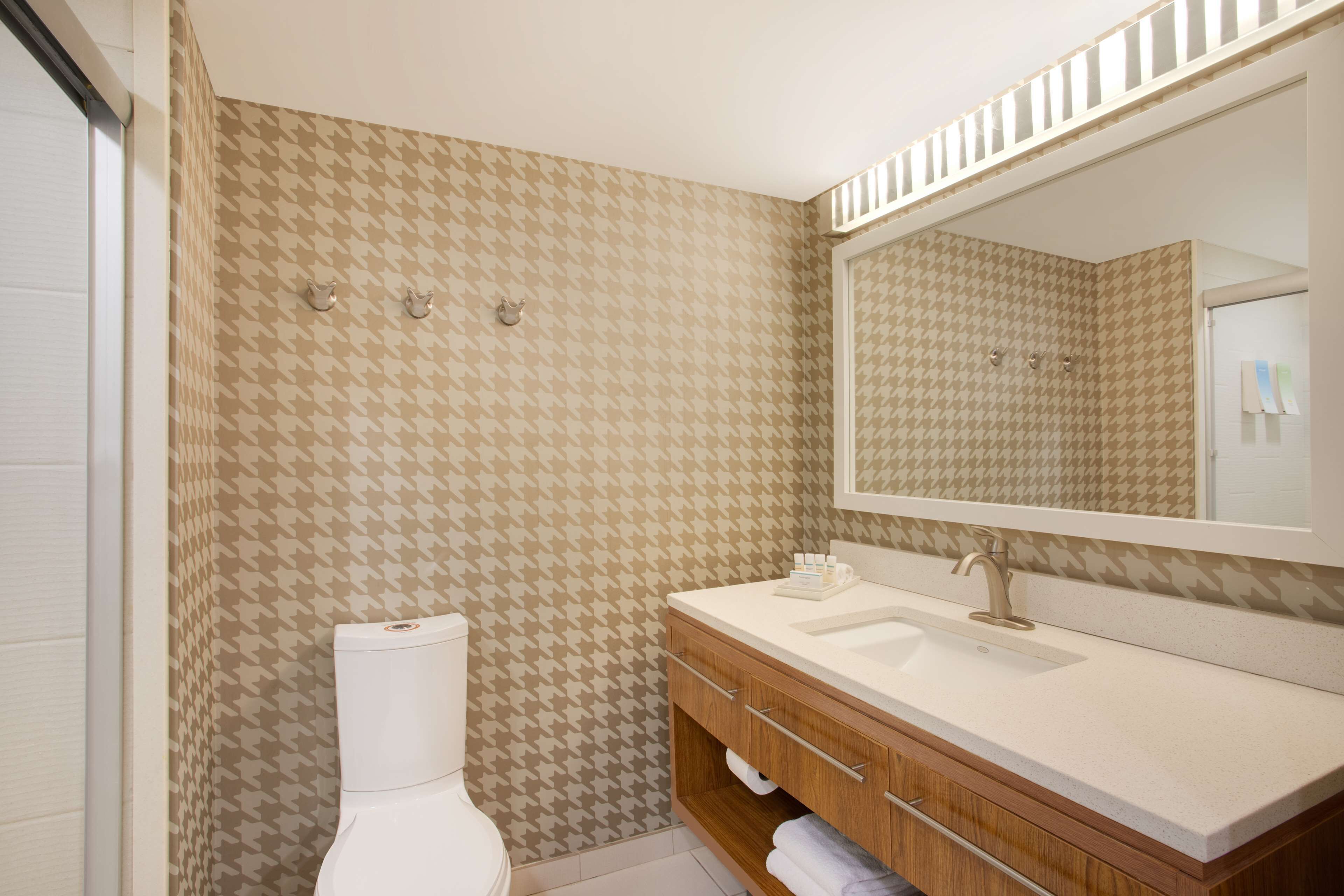 Une salle de bain avec un mur à motifs, des toilettes blanches, un meuble-lavabo en bois avec un miroir et un évier.