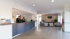 Un espace d’accueil comprenant un comptoir, deux employés, une horloge, un téléviseur fixé au mur et un coin salon agrémenté d’une plante.