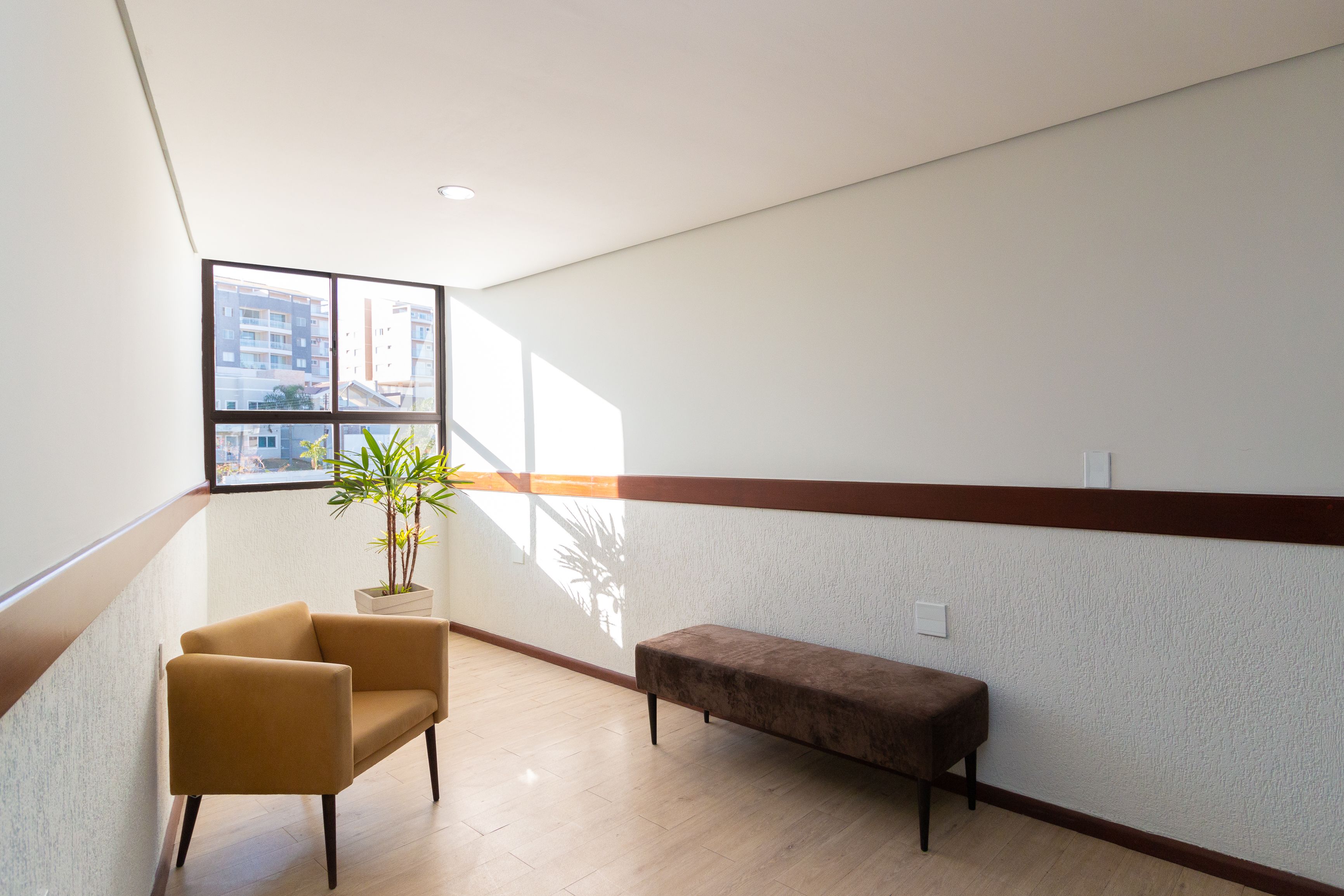 A modern interior with a wooden floor, a brown bench, a yellow armchair, and a potted plant by a window with a view of buildings.