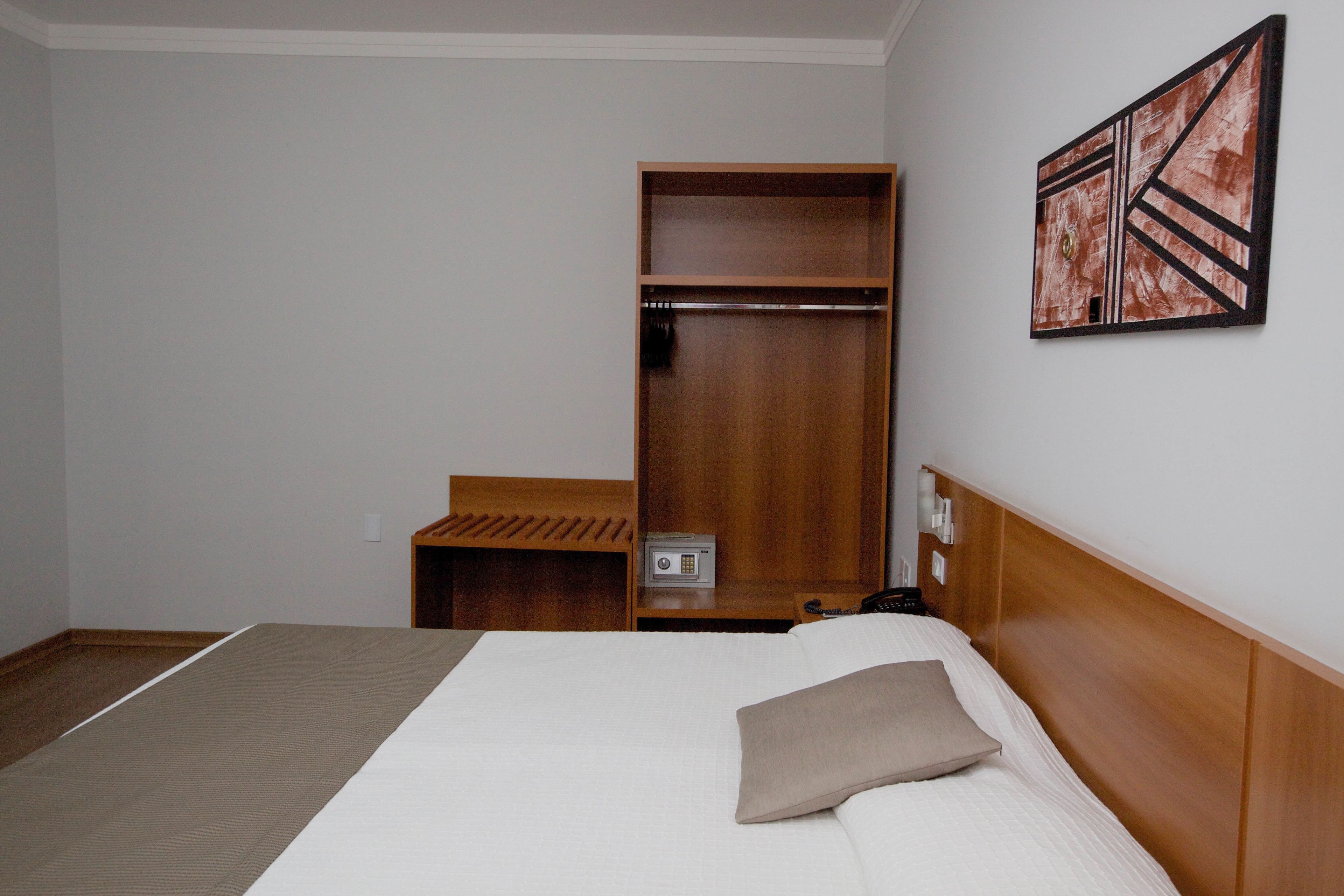 A hotel room with a bed, a wooden wardrobe, a bedside table, and a wall-mounted artwork.