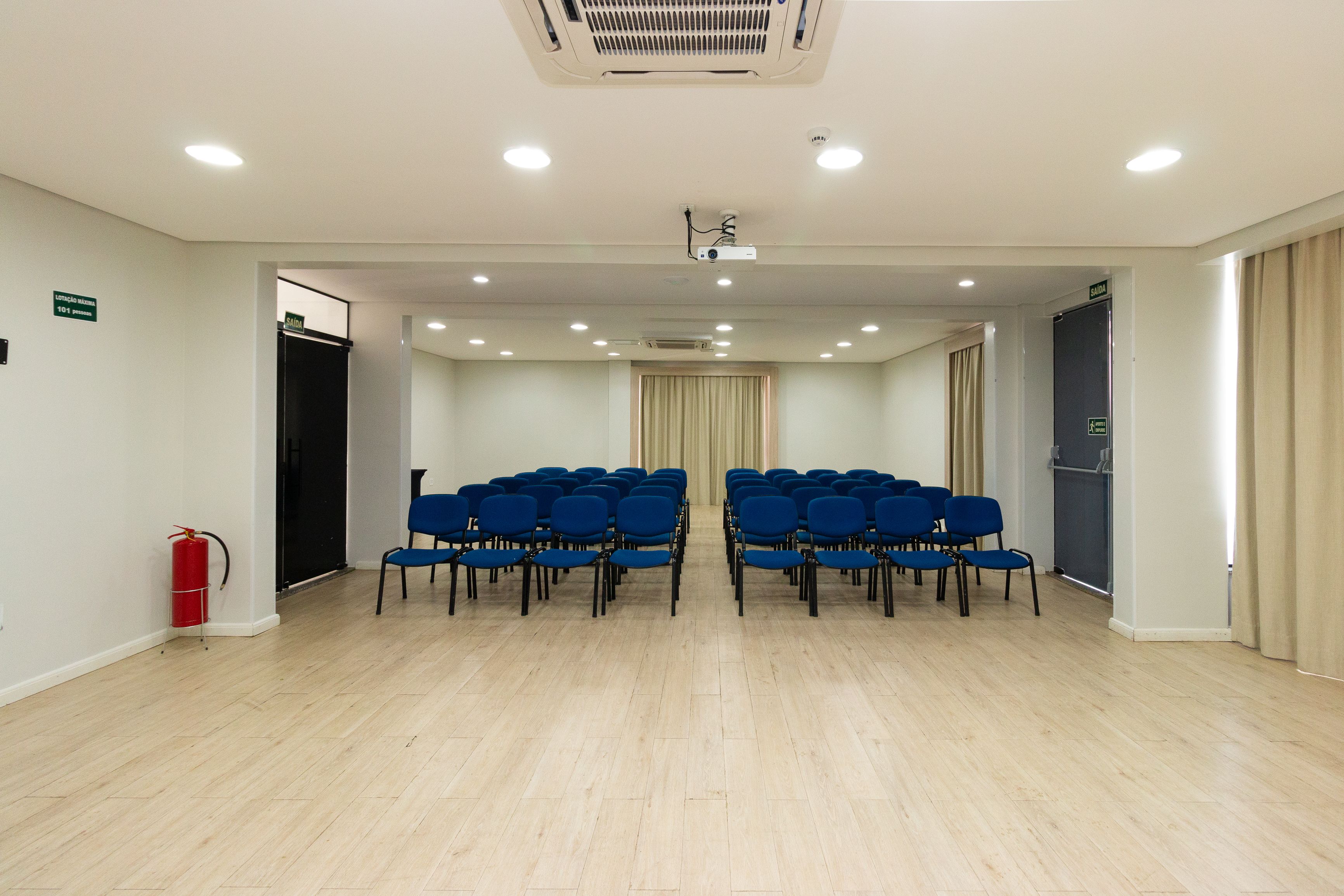 A conference room with rows of blue chairs, a fire extinguisher, and a projector on the ceiling.