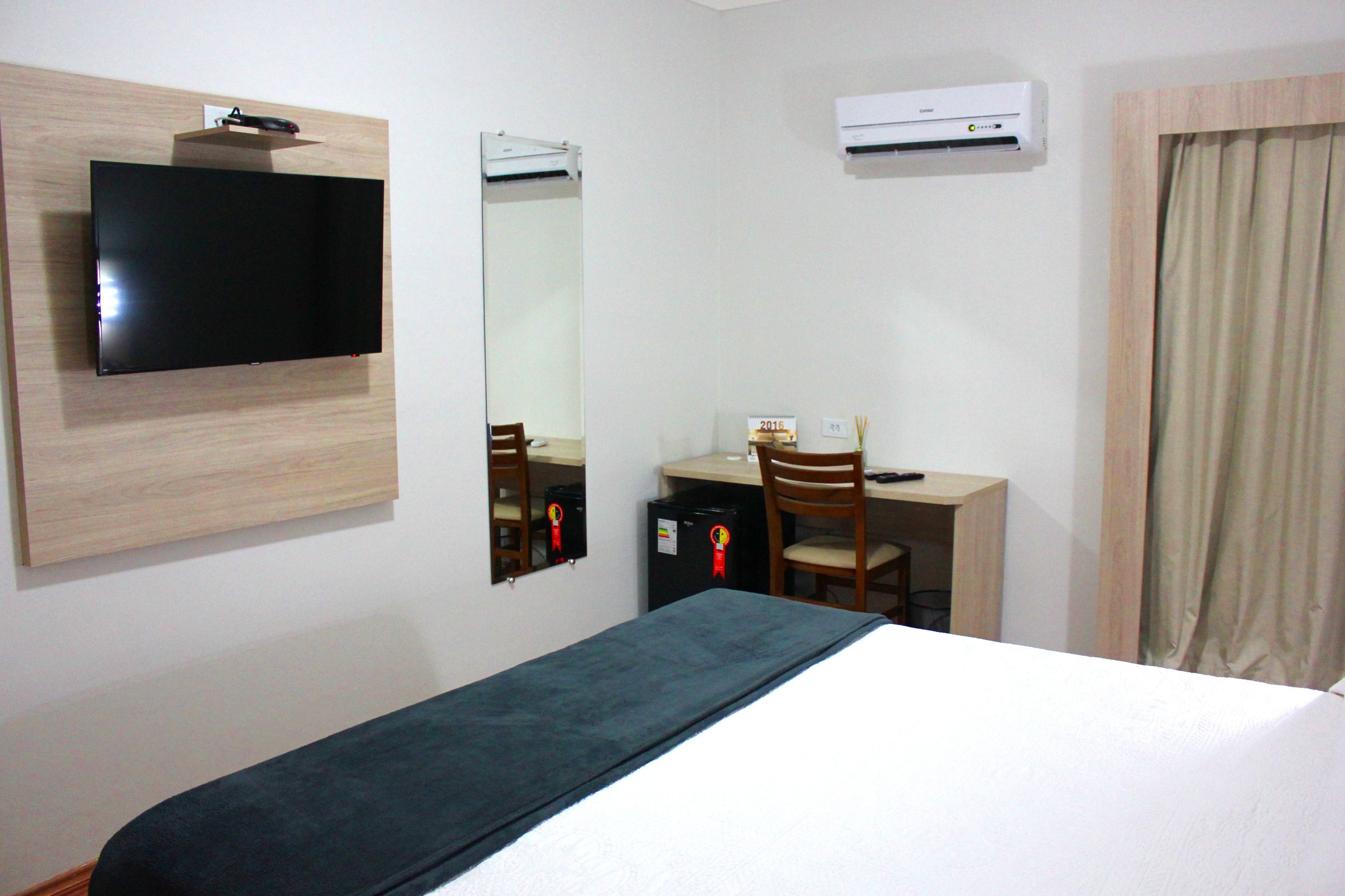 A hotel room with a bed, a desk with a chair, a television, a mirror, and an air conditioning unit.