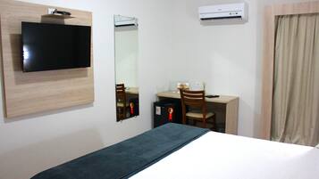 A hotel room with a bed, a desk with a chair, a television, a mirror, and an air conditioning unit.