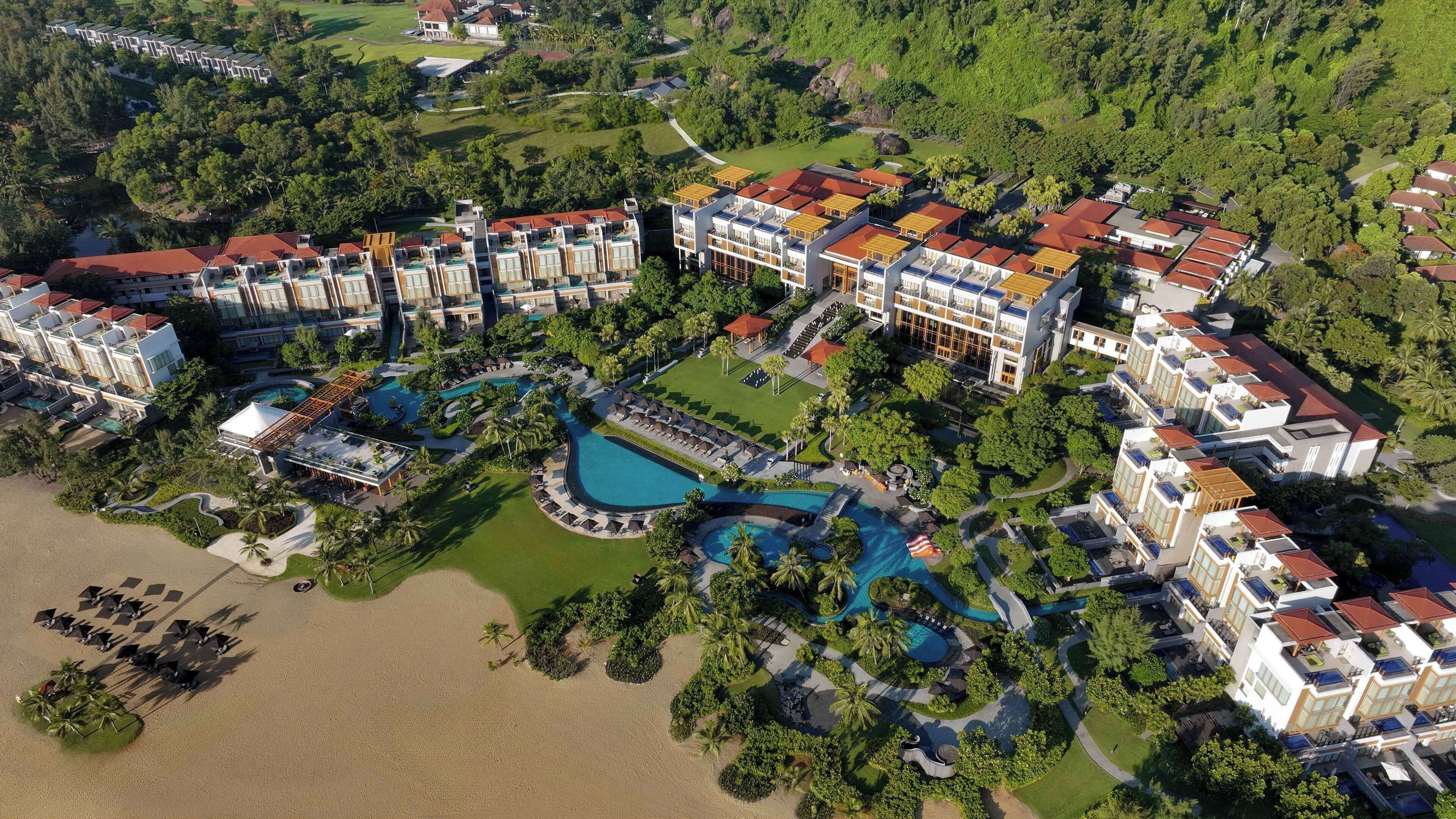 An aerial view of a resort with multiple buildings, a large central pool, and surrounding greenery.