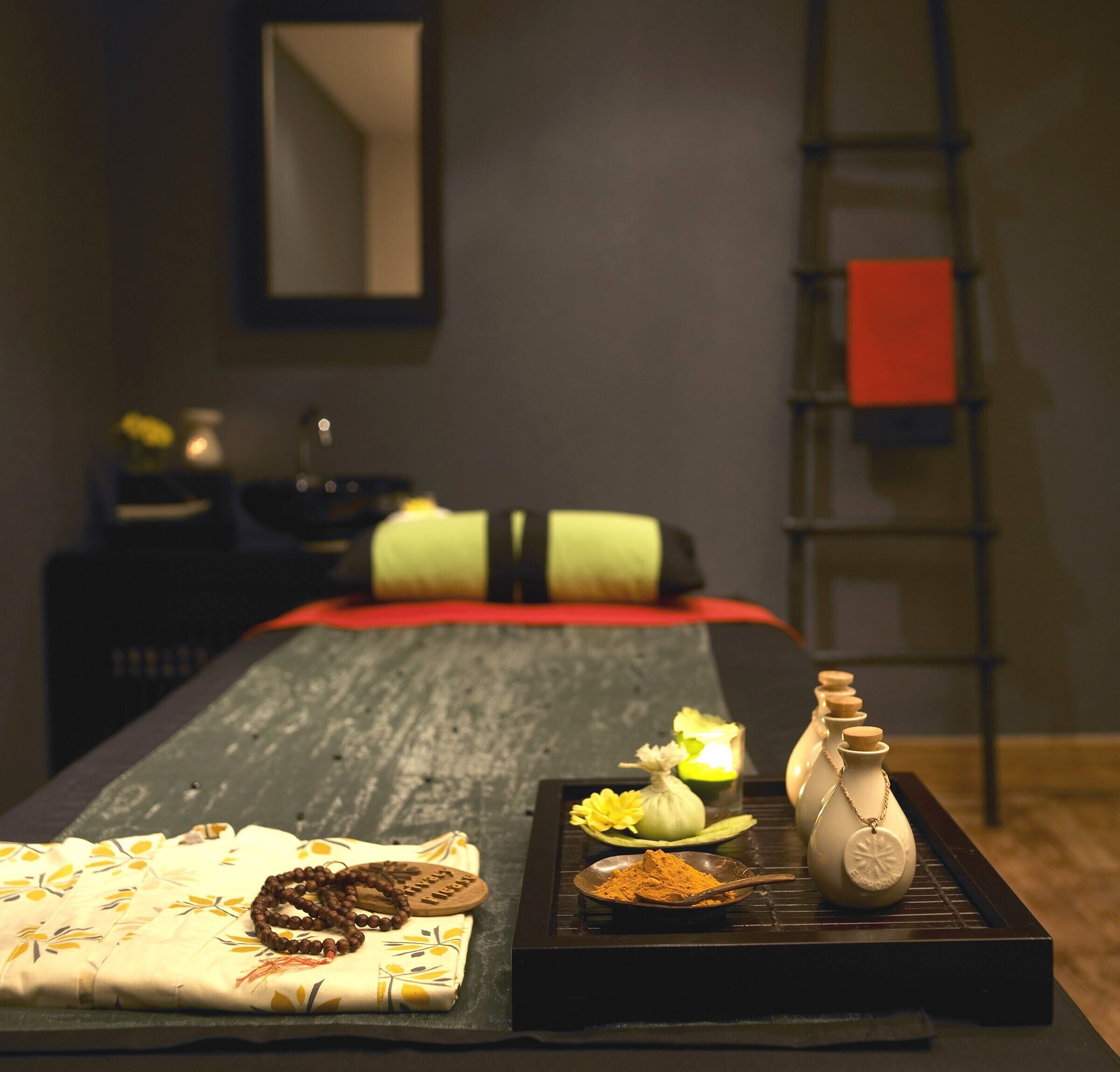 A neatly arranged massage table with a floral bedspread, a wooden tray with a beaded necklace, a small bowl of yellow powder, and a white vase with a cork stopper.