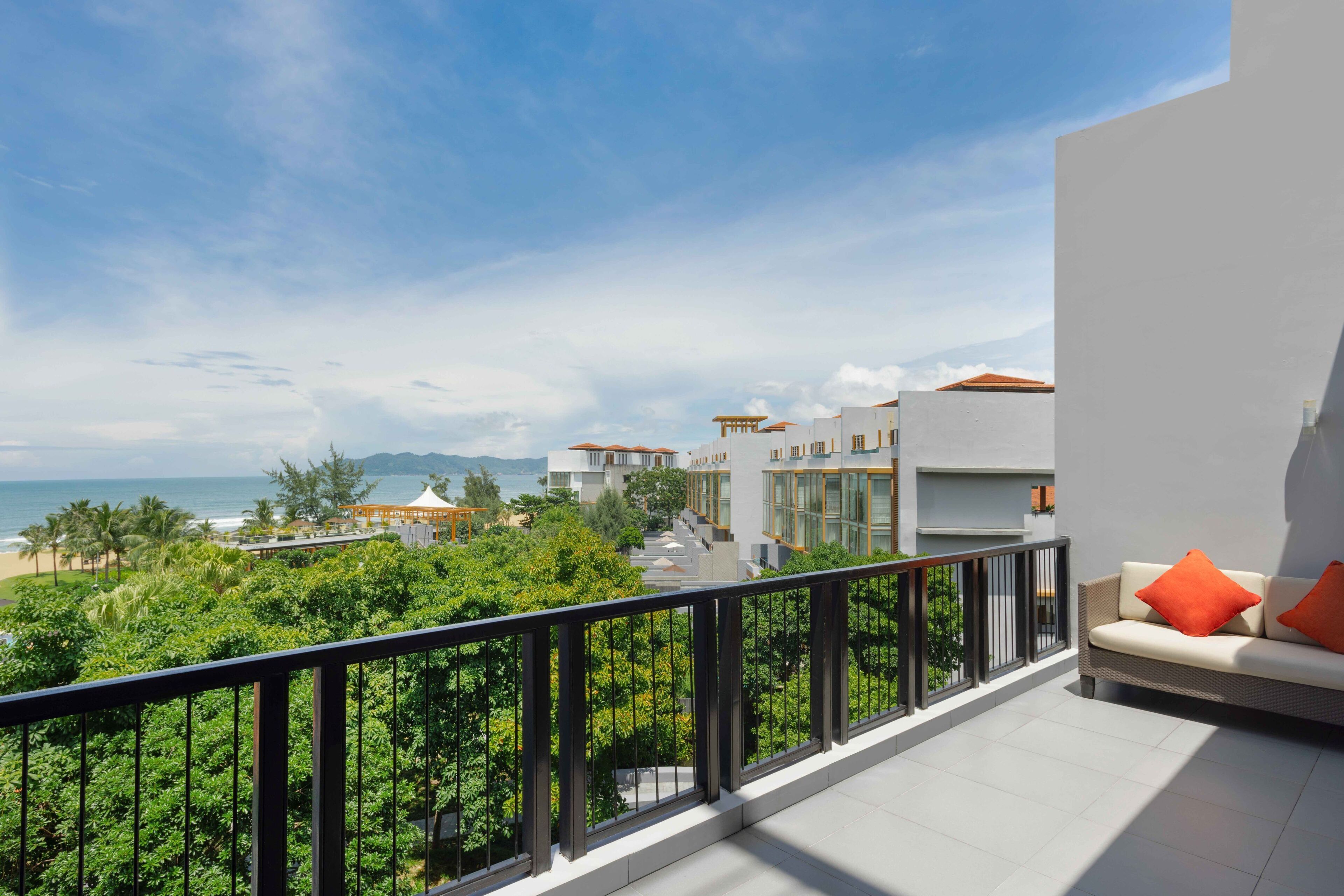 A balcony with a view of the ocean and greenery, a white wall, and a sofa with orange cushions.