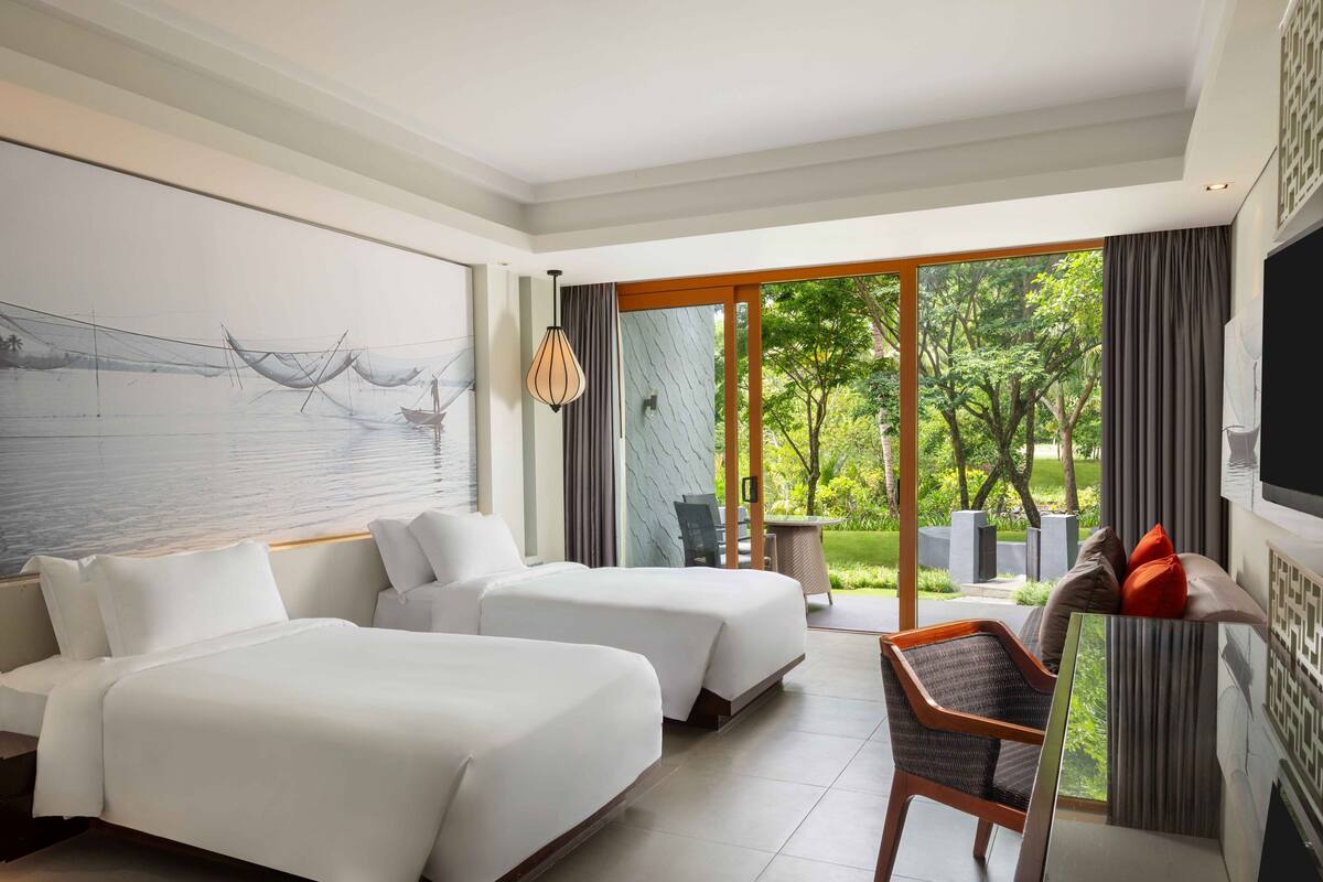 A hotel room with two beds, a seating area with a chair, a large window with a view of greenery, and a wall-mounted TV.